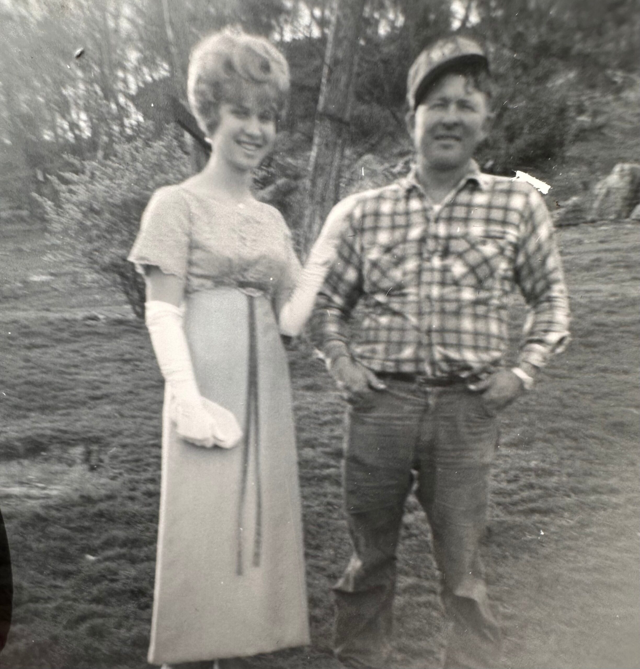 Two people stand outdoors with a tall rectangular wooden post between them; a smiling woman in a light dress on the left and a man in a plaid shirt and cap on the right, with trees in the background.