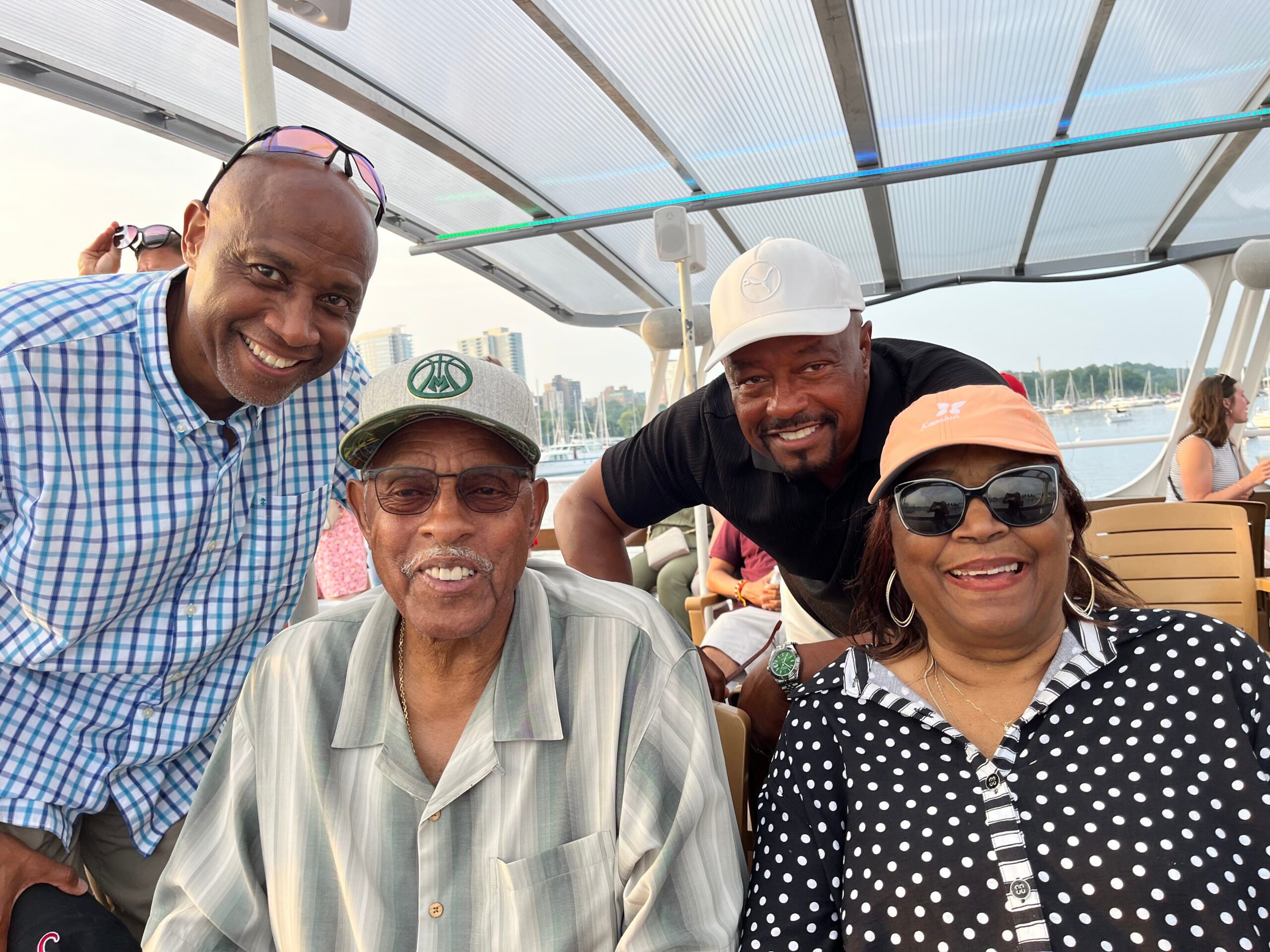 Group of smiling adults taking a boat selfie with a marina and city skyline in the background