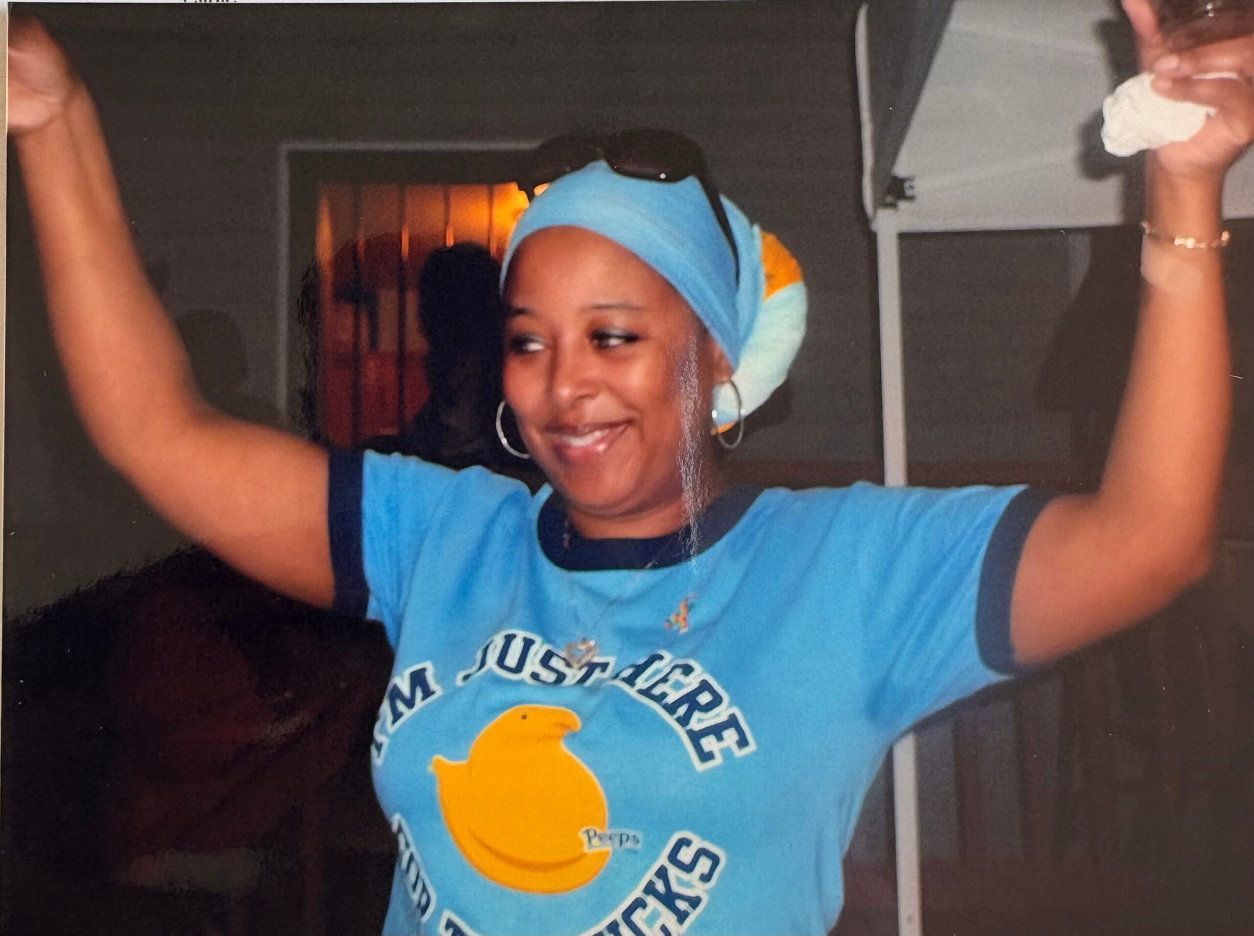 Woman dancing with arms raised, wearing a blue head wrap and blue T‑shirt, smiling at a party or social event.