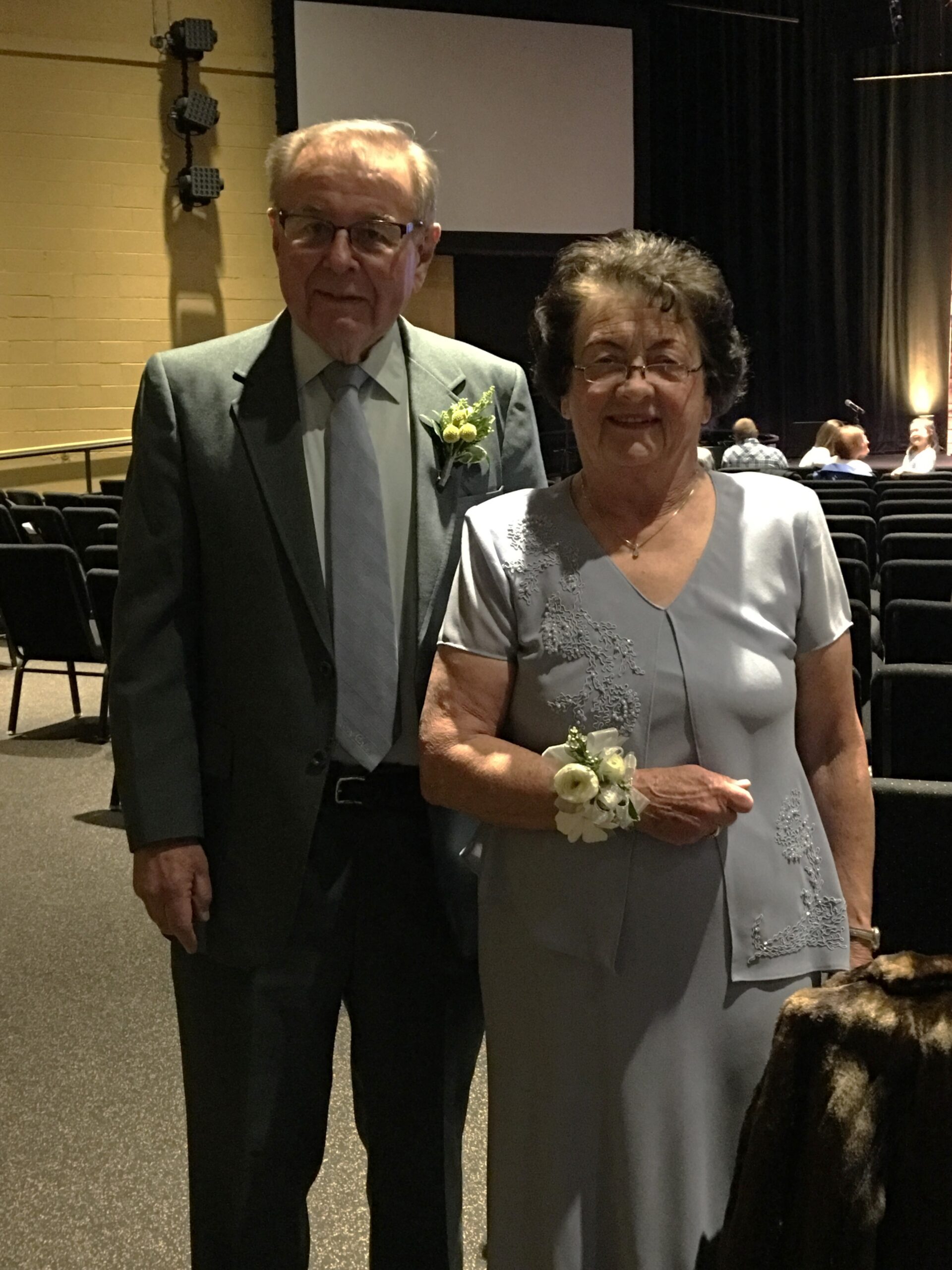 An elderly man in a gray suit and tie with a yellow corsage standing beside an elderly woman in a gray dress with floral embroidery inside a dim auditorium.