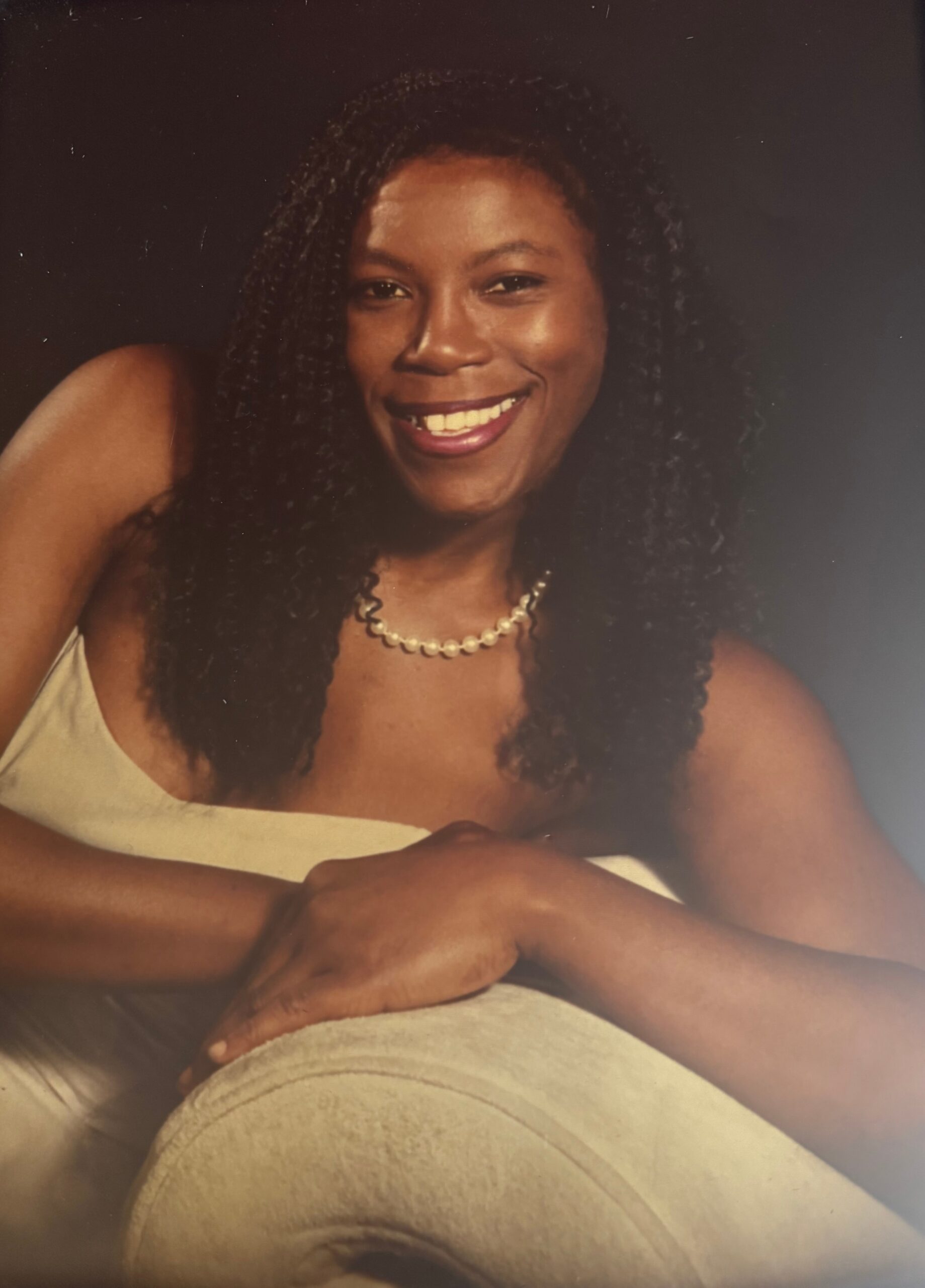 Smiling woman with long curly hair wearing a pearl necklace and off-shoulder light dress against a dark backdrop—portrait style photo