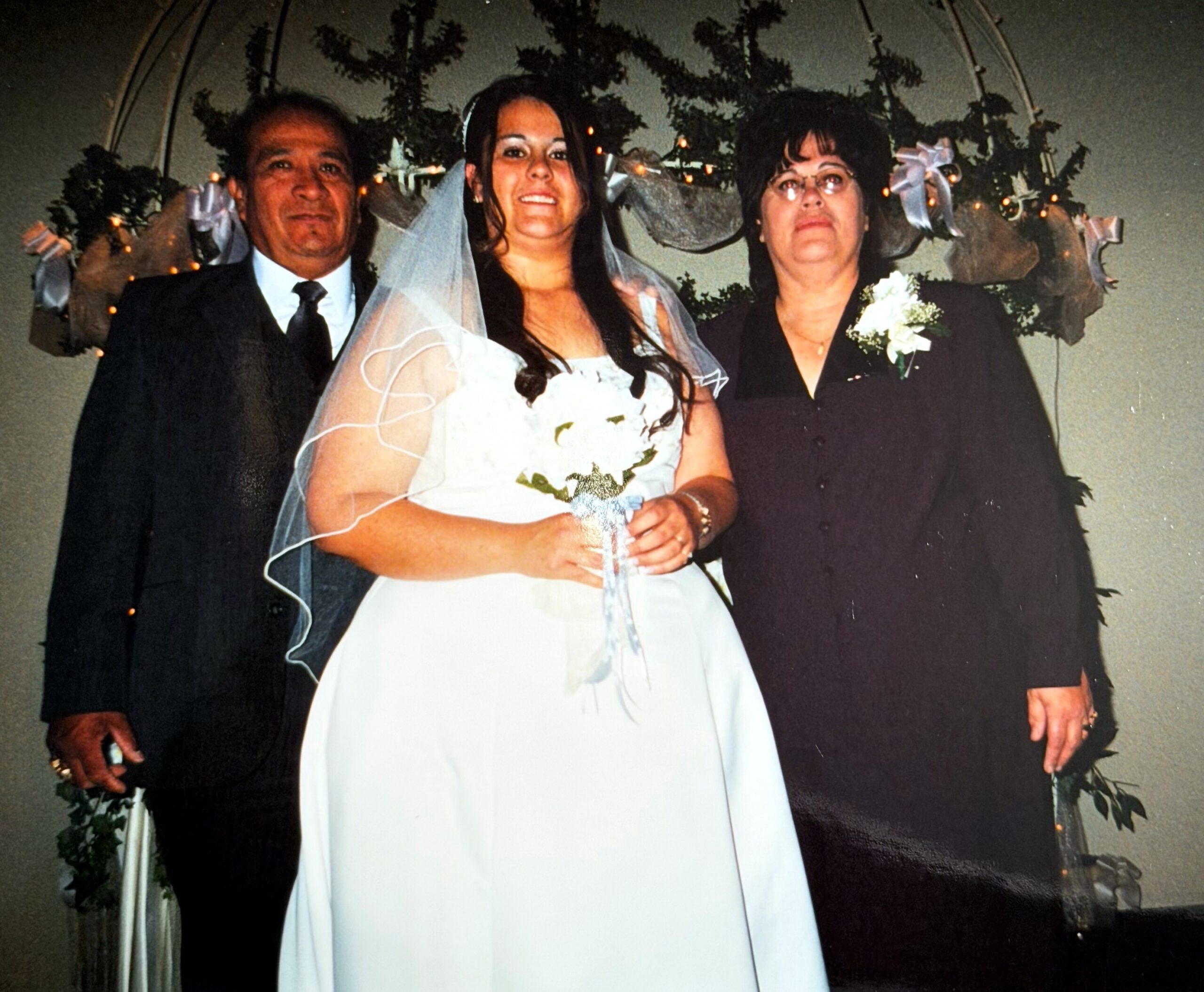 Bride in a white wedding dress with veil, standing between a man and a woman in formal black attire at a wedding.