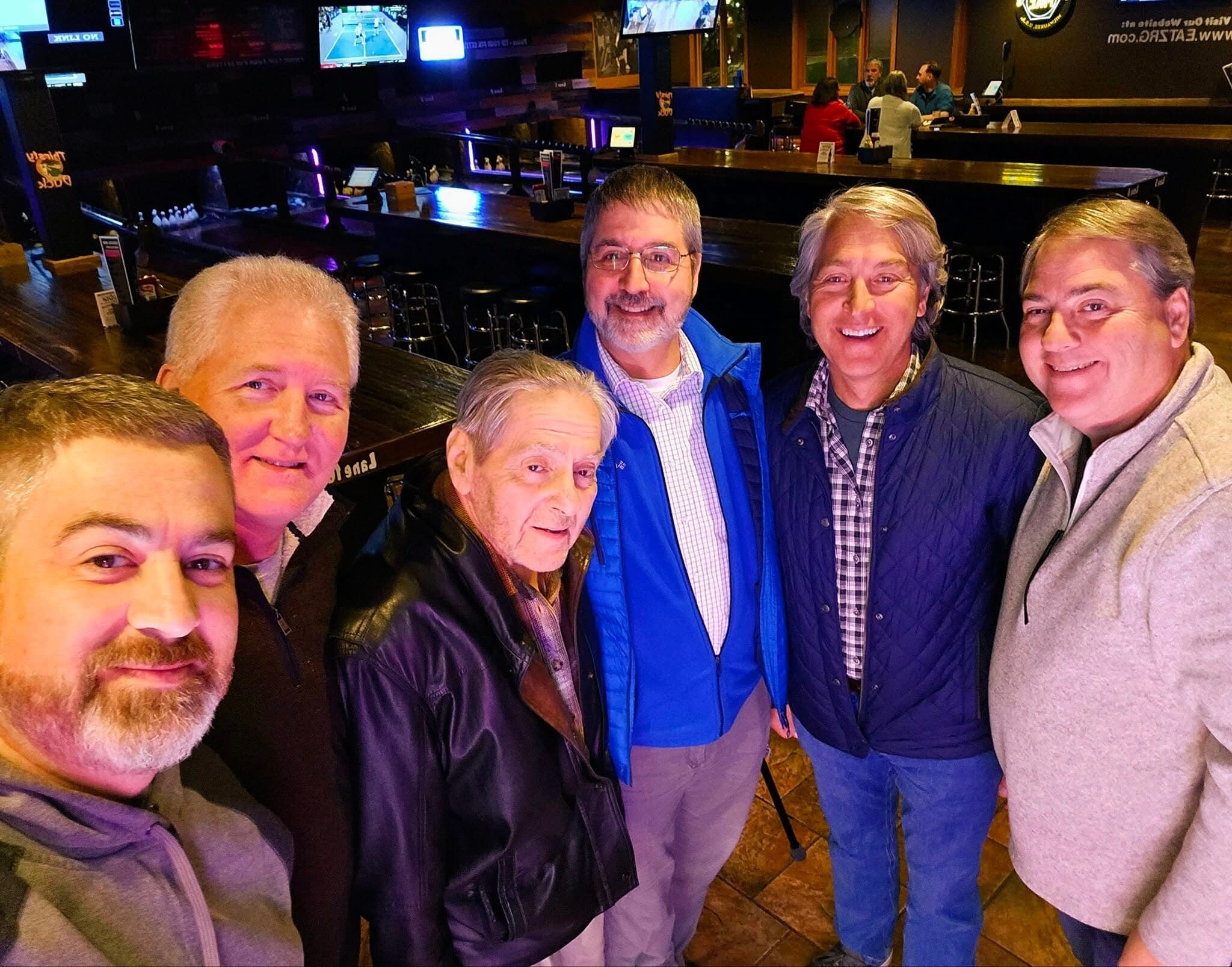 Six men take a casual group selfie at a sports bar, wearing jackets, with TVs and a dark bar backdrop behind them.