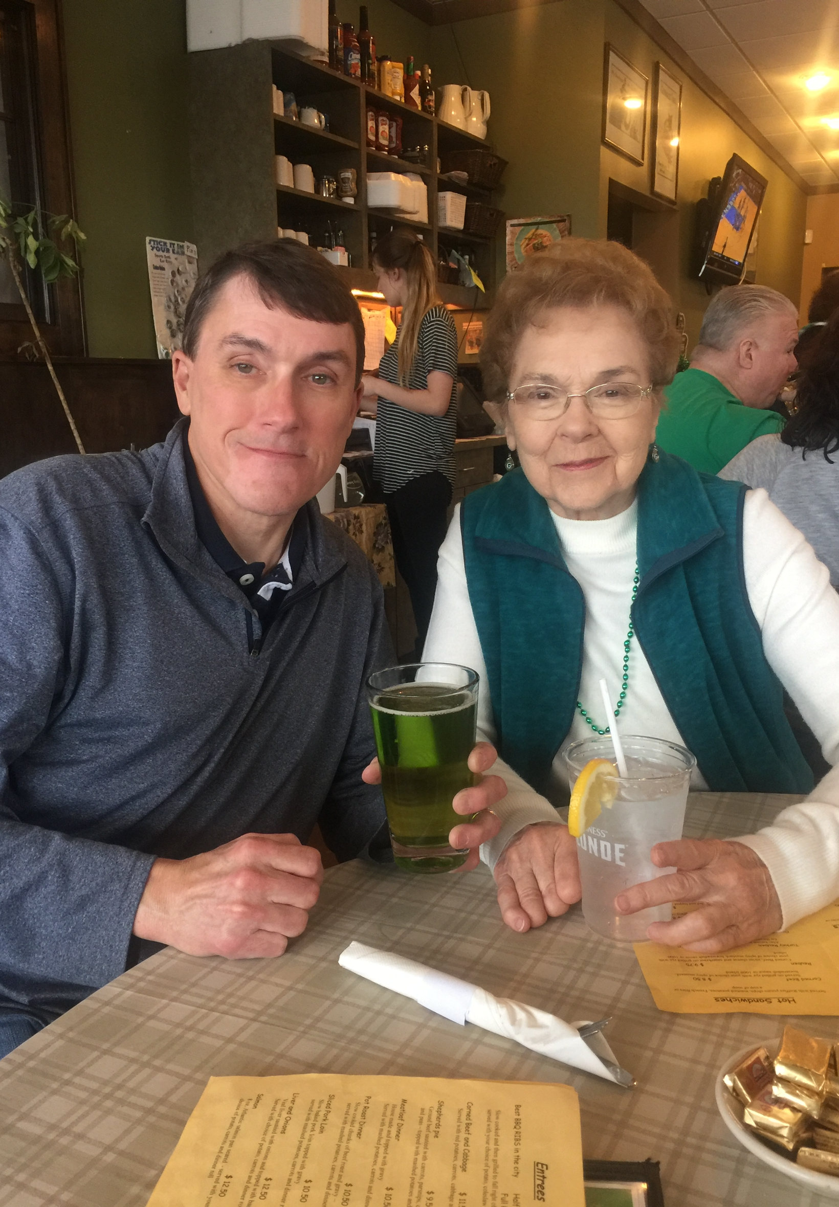 Two people sit at a restaurant table with drinks; man holding a green beverage, older woman in teal vest and glasses beside him, smiling at camera .