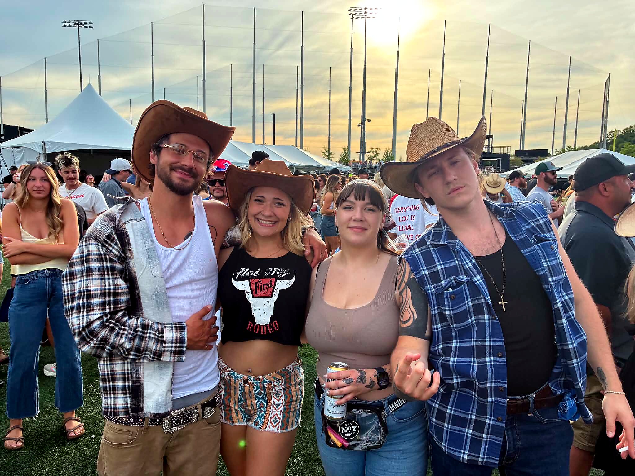 Group of friends at an outdoor festival posing for a photo, several wearing cowboy hats with tents and light poles in the background.