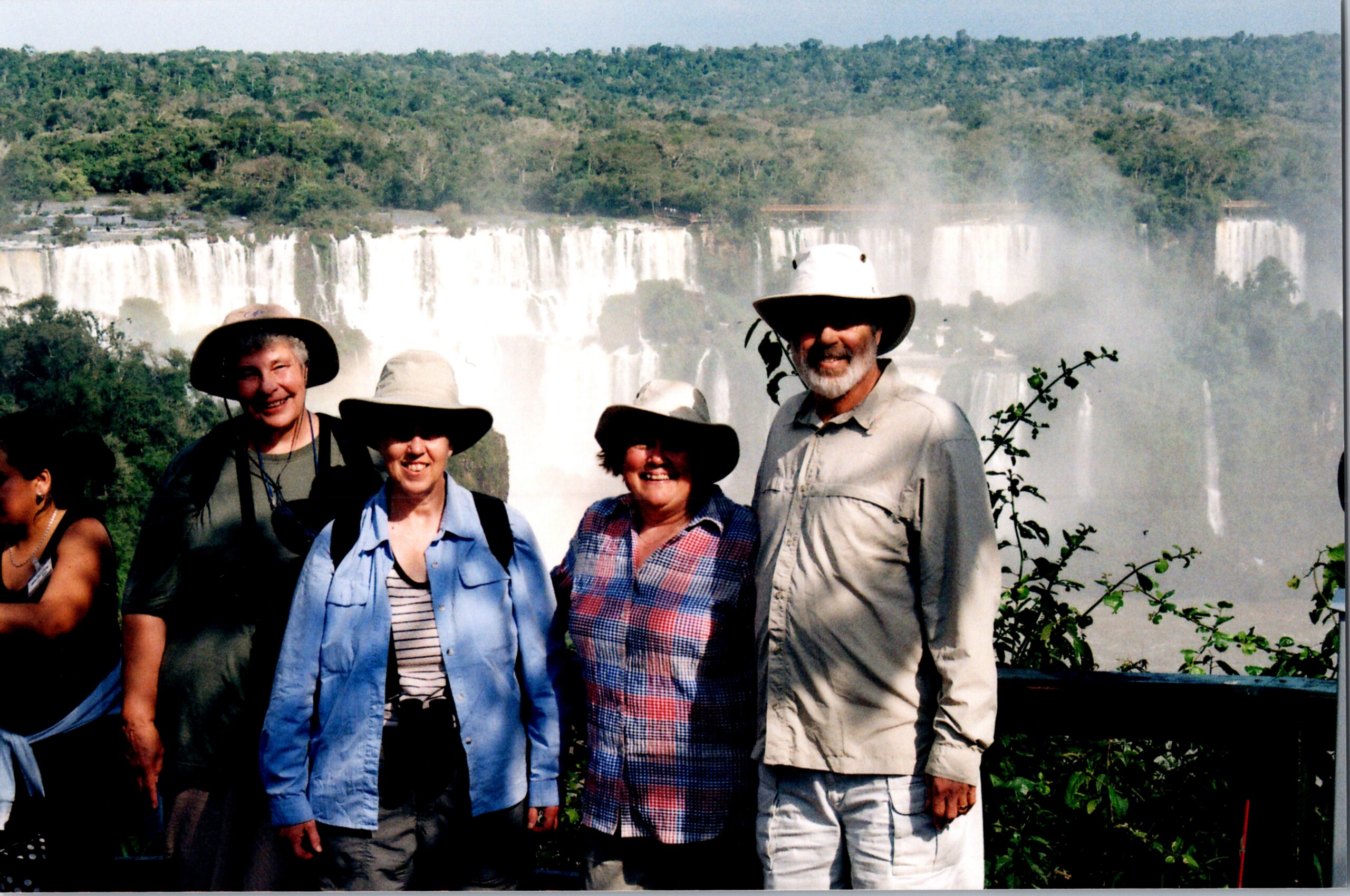Smiling group of tourists wearing hats posing in front of a large waterfall with mist and green forest in the background.