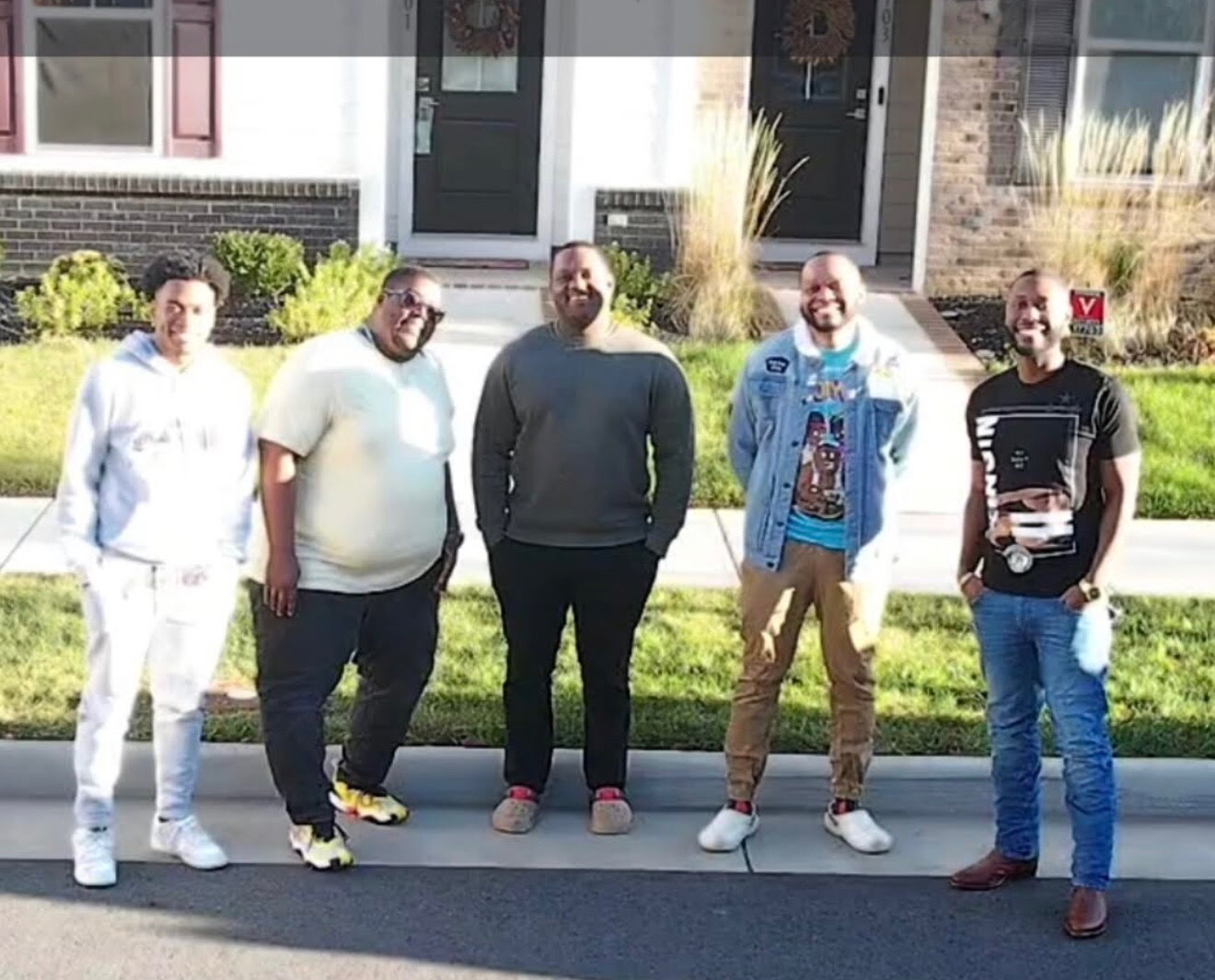 Five men stand in a row on a sidewalk in front of brick townhouses, smiling at the camera.
