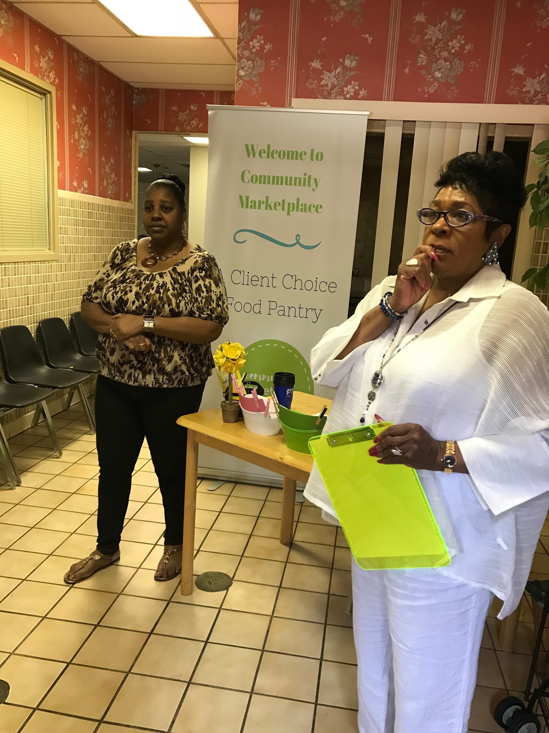 Two women stand near a banner that reads 'Welcome to Community Marketplace Client Choice Food Pantry' in a tiled room.