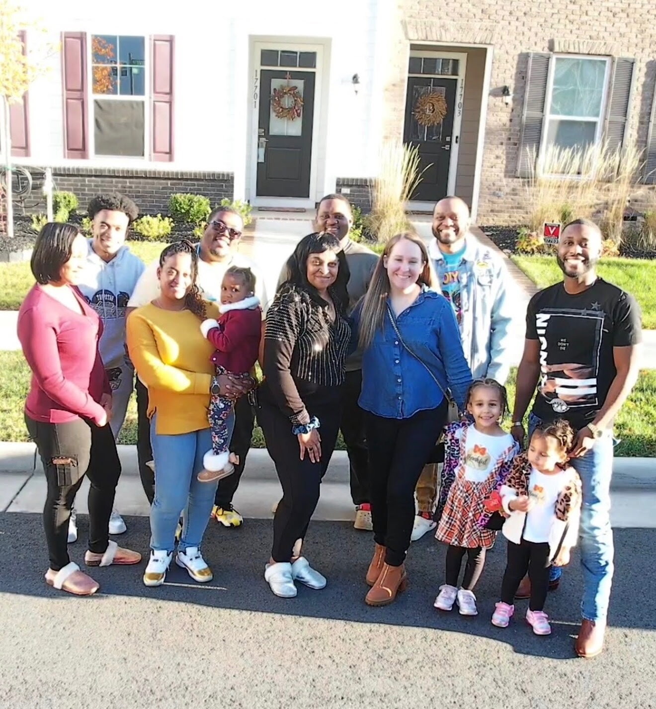 Group of adults and children posing together on a sunny suburban sidewalk in front of brick houses with wreaths on the doors, smiling at the camera.