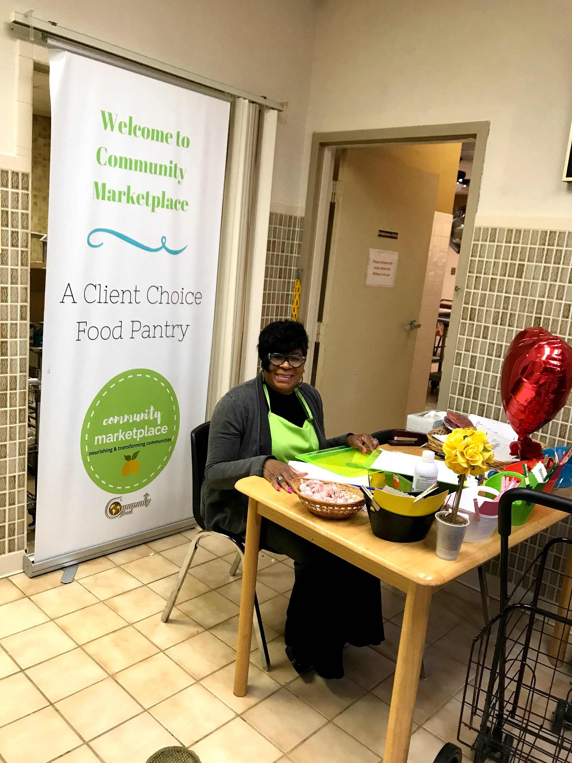 Smiling woman in a green apron sits at a craft table next to a 'Welcome to Community Marketplace' banner.