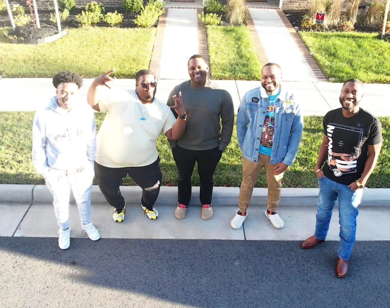 Five men stand in a row on a curb, posing and smiling for a casual group photo in a suburban neighborhood.