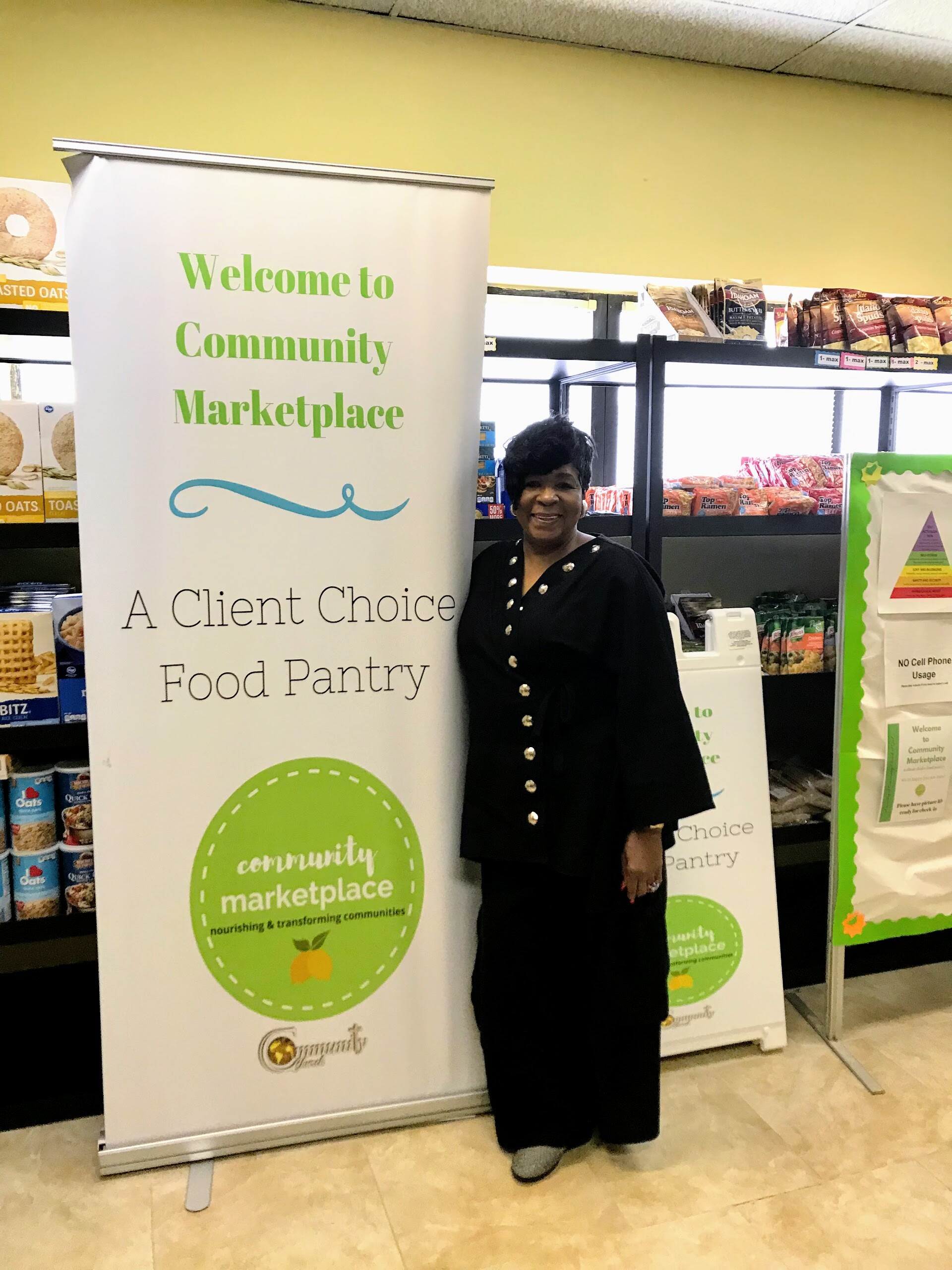 Smiling woman in a black outfit stands beside a banner reading 'Welcome to Community Marketplace — A Client Choice Food Pantry' in a store.