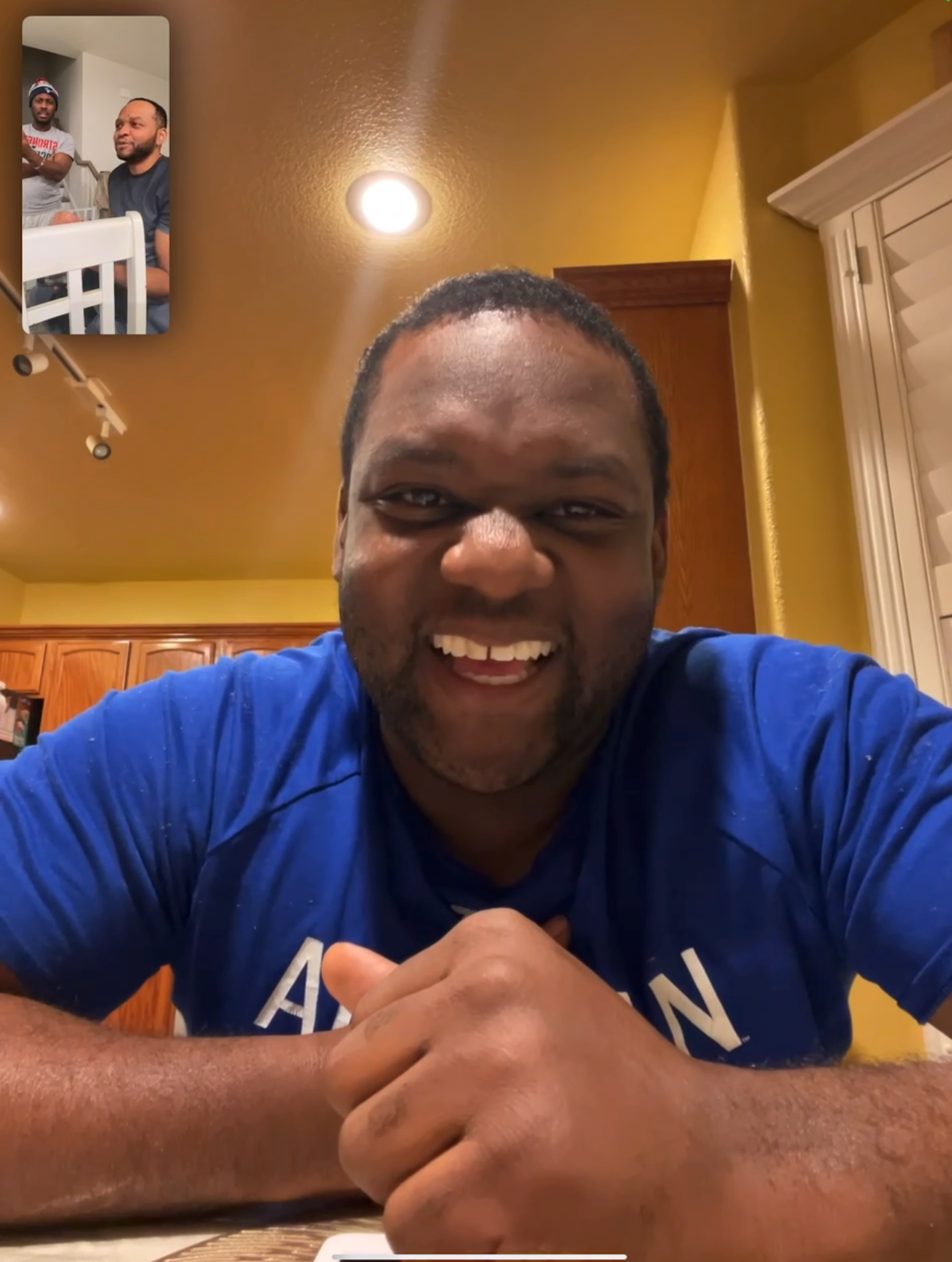 Smiling man in a blue shirt on a video call, kitchen background; inset shows two men chatting in another location.