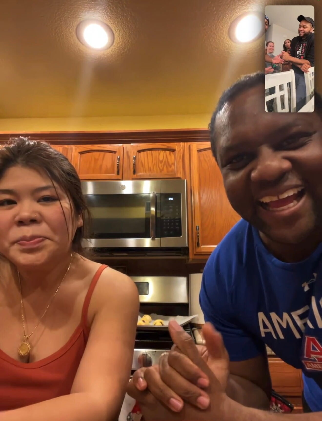 Two people smiling in a kitchen during a video call, with wooden cabinets and a stainless oven in the background.