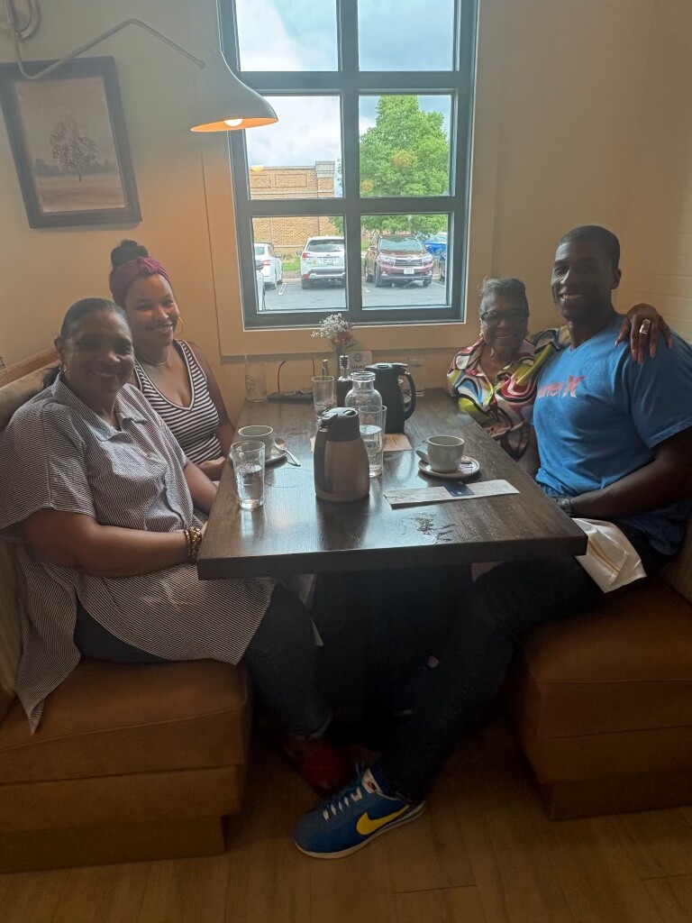 Four people sitting around a table in a cafe, smiling at the camera with coffee cups and a carafe on the table.