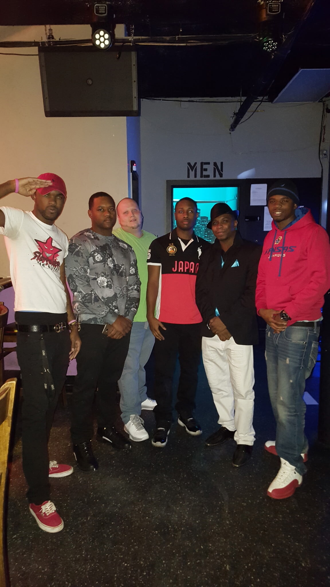 Six men posing together indoors in a venue, standing in a row in front of a wall that says MEN; one man salutes the camera on the far left.