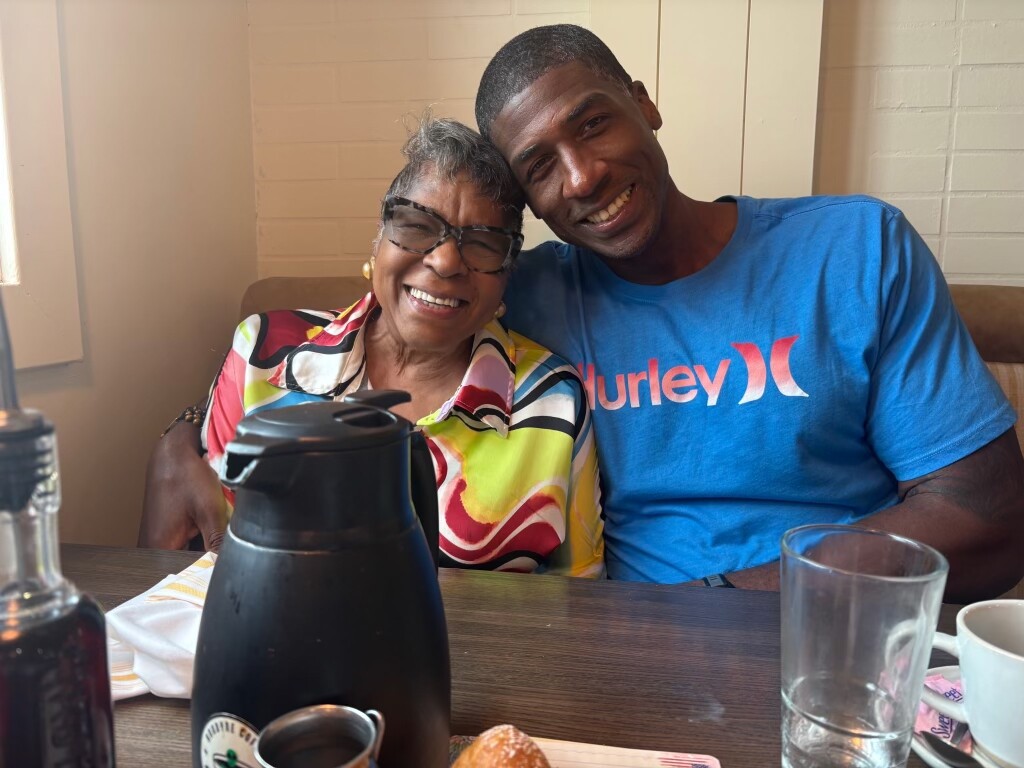 Two people sit close together at a restaurant table, smiling warmly at the camera; a black coffee carafe and glass are on the table in front of them. The man wears a blue Hurley shirt and the woman wears a colorful patterned blouse and glasses.