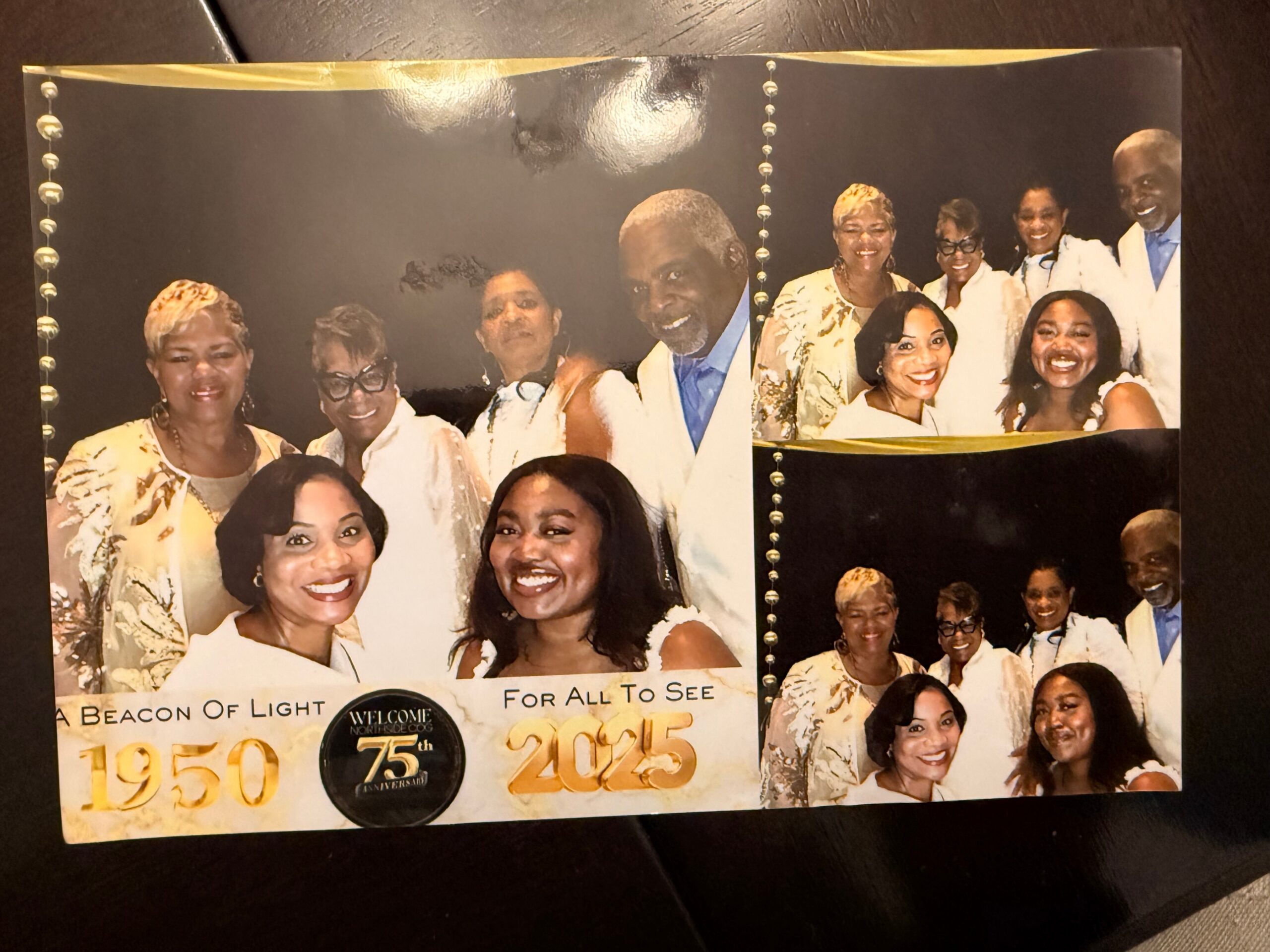 Group portrait of smiling adults in white attire posing together at a celebratory event, with a dark backdrop and bead decorations behind them.