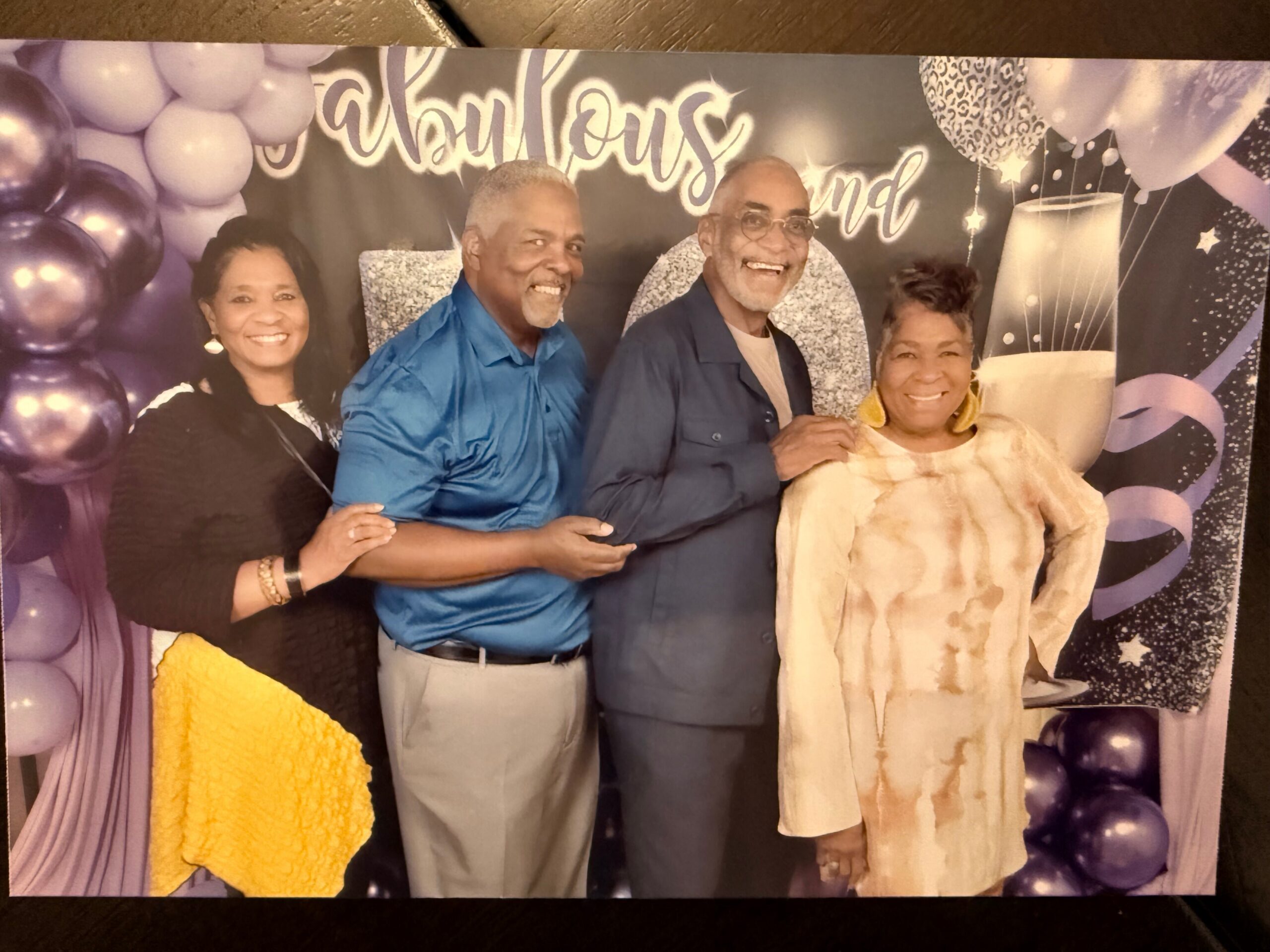 Four smiling adults posing together in front of a festive party backdrop with purple balloons and a champagne glass design on the banner.