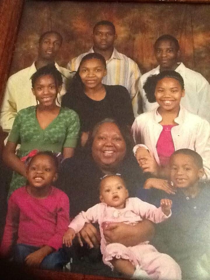 A smiling multigenerational African‑American family posing for a studio portrait, with a woman in black holding a baby in pink at center and relatives gathered around.