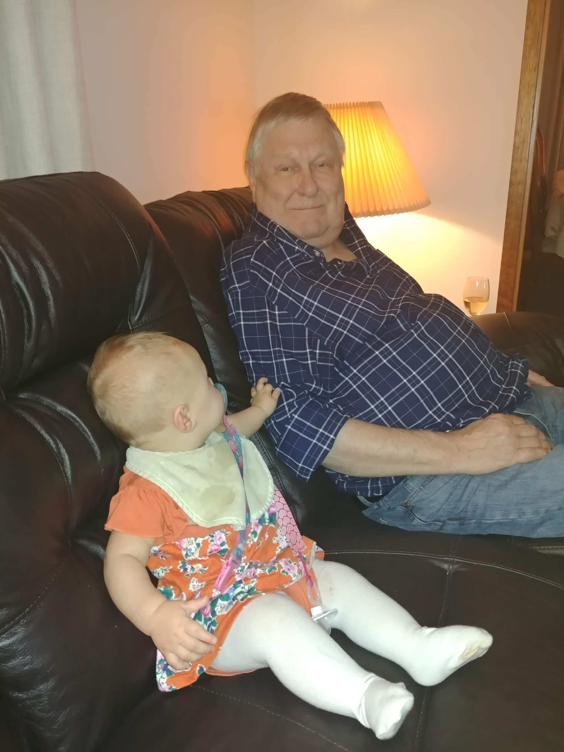 Older man in a plaid shirt sits on a brown leather couch with a baby in a floral dress beside him, warm lamp behind them.