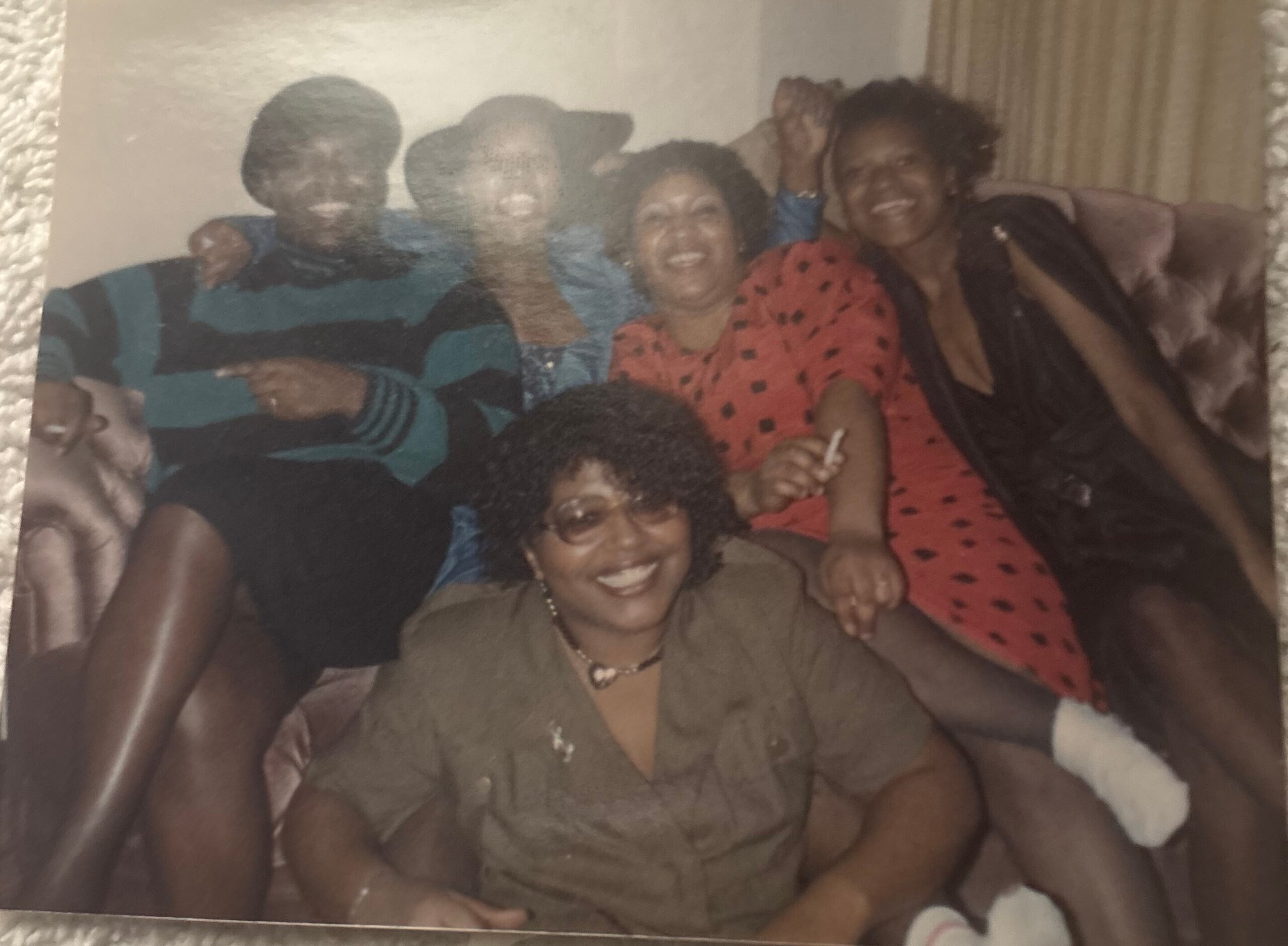 Group of friends smiling and posing together on a couch in a living room, wearing casual colorful clothing.