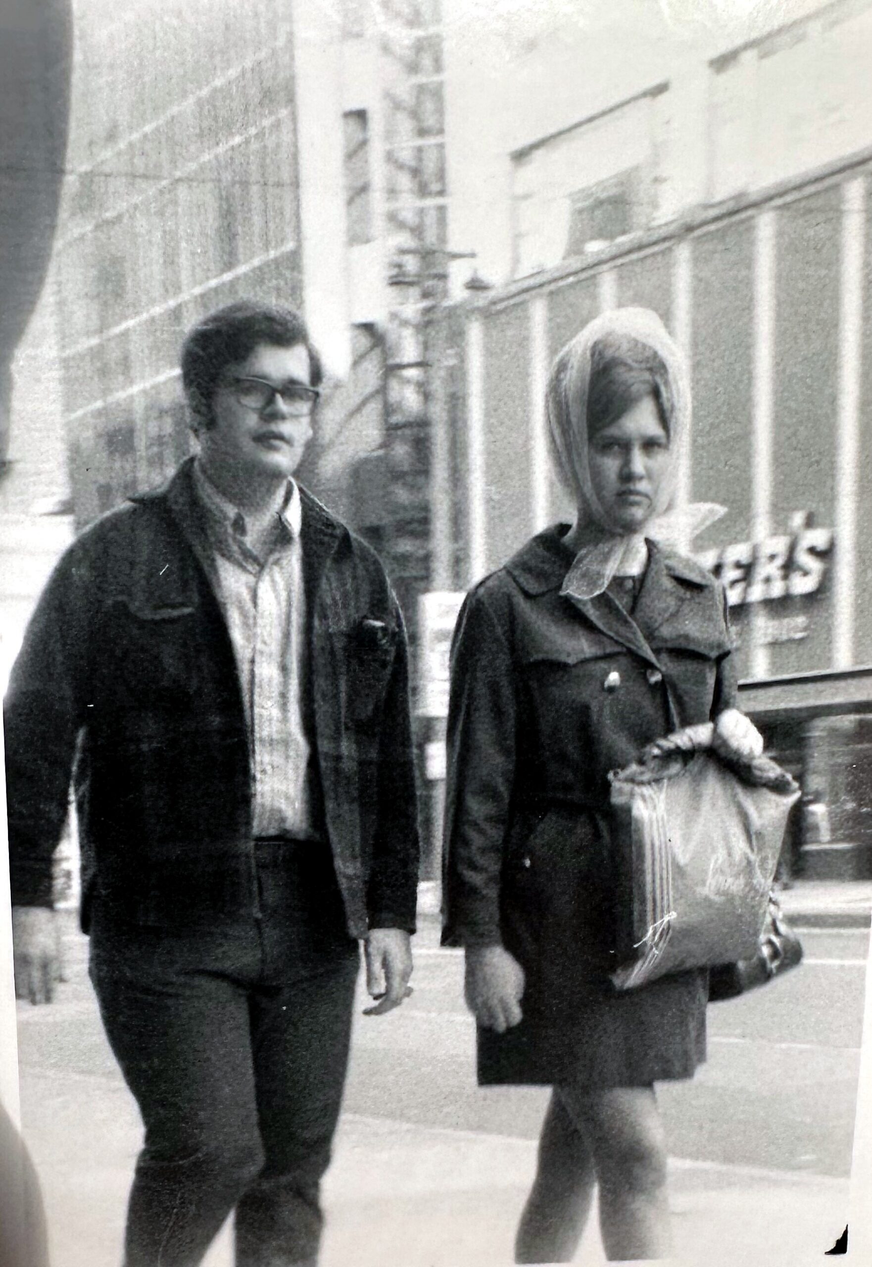 Man with glasses walking beside a woman wearing a headscarf on a city sidewalk.