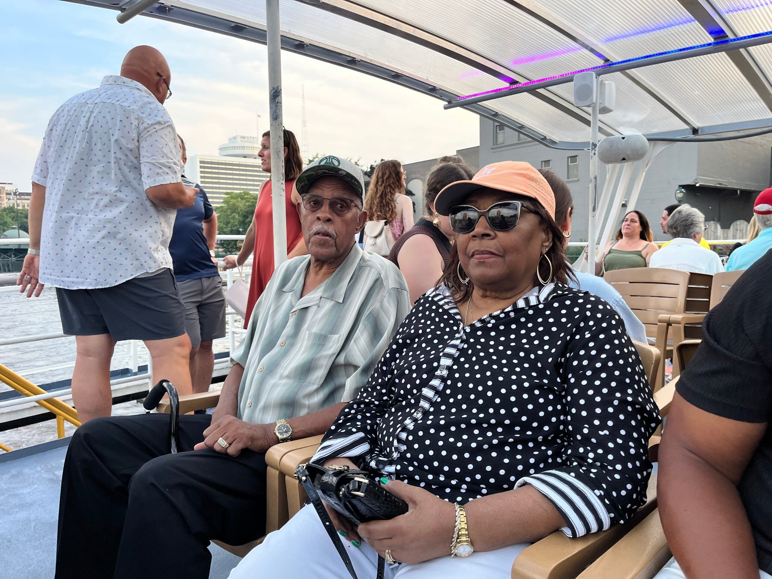 Adults seated on a boat deck, woman in a black-and-white polka-dot blouse wearing sunglasses in the foreground and a man in a light striped shirt nearby, with city buildings in the background and a translucent roof above.