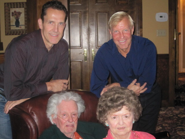 Four adults pose for a family photo in a living room: two men stand behind an elderly couple seated in a leather chair, all smiling.