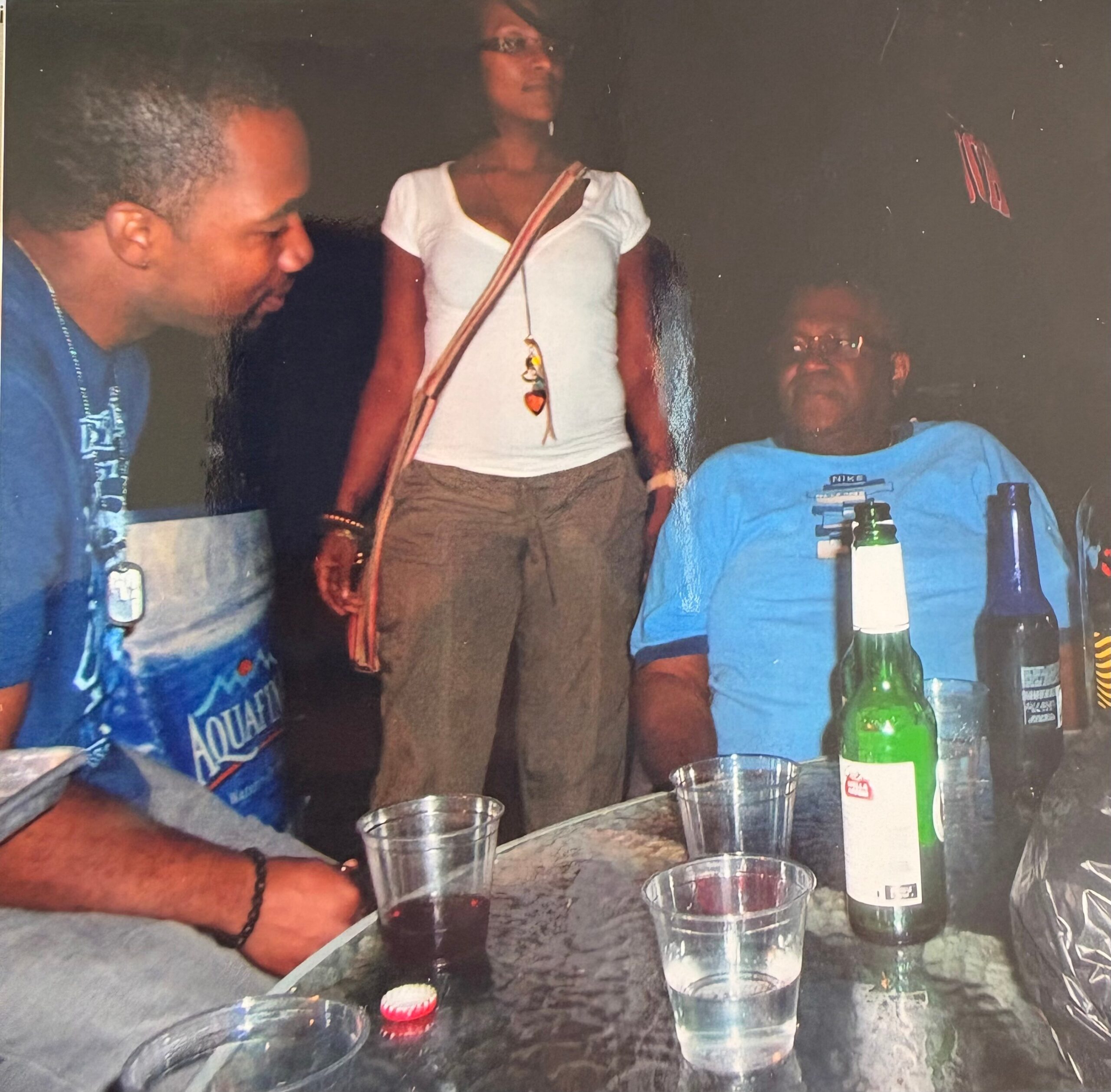 Three people sit and stand around a glass table with beer bottles and plastic cups; a woman in a white top stands in the center wearing a striped bag strap across her chest.
