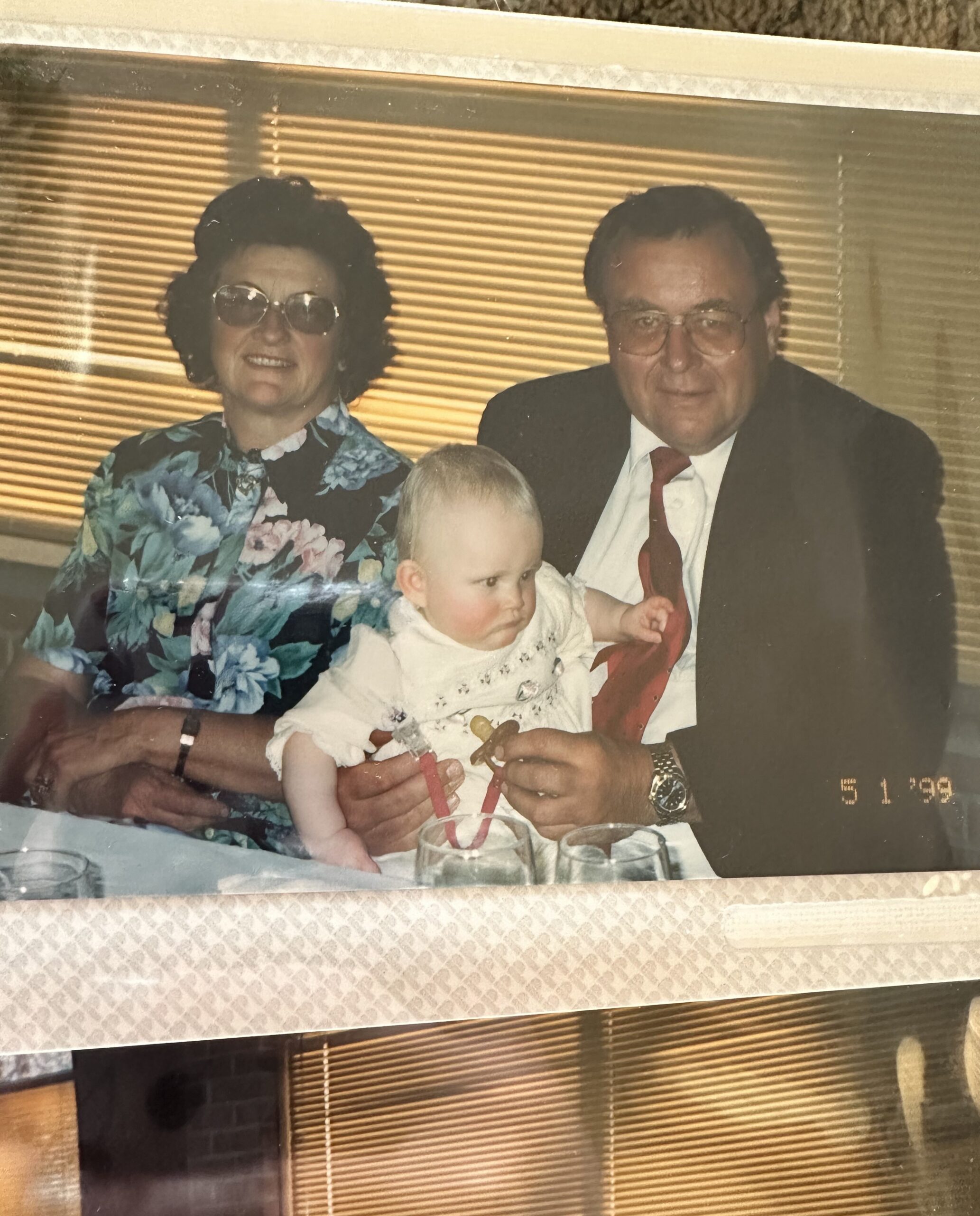 Woman in a floral dress holds a baby while a man in a suit sits beside them, against striped blinds in the background.