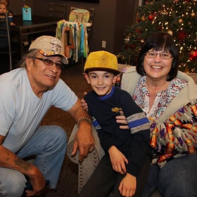 Three people pose for a holiday photo on a living room carpet: an older man in a light shirt and cap, a smiling boy in a yellow hat, and an older woman in a patterned cardigan, with a Christmas tree in the background.