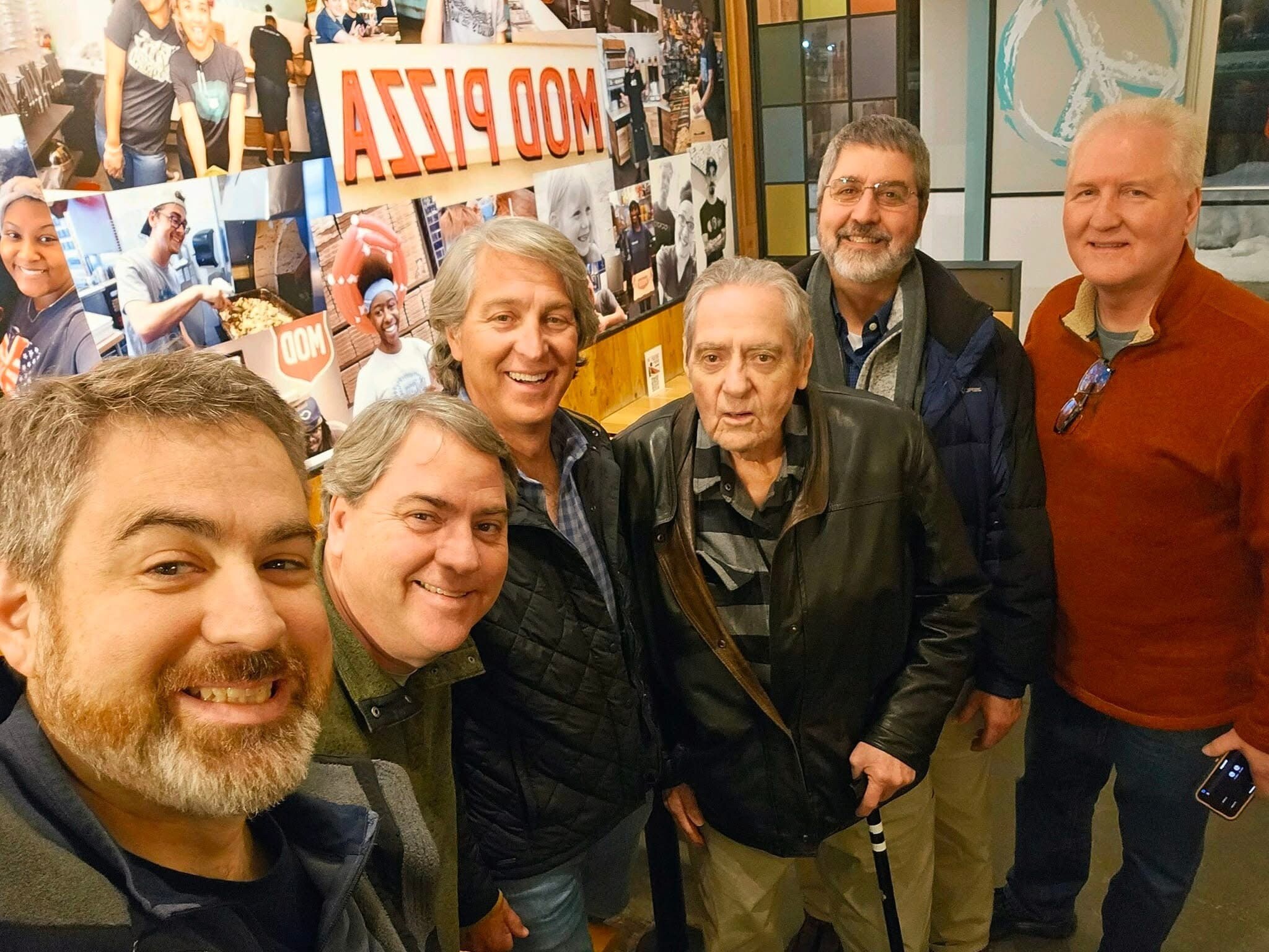 Group of six adults taking a close-up selfie indoors, smiling, with a photo collage wall in the background.