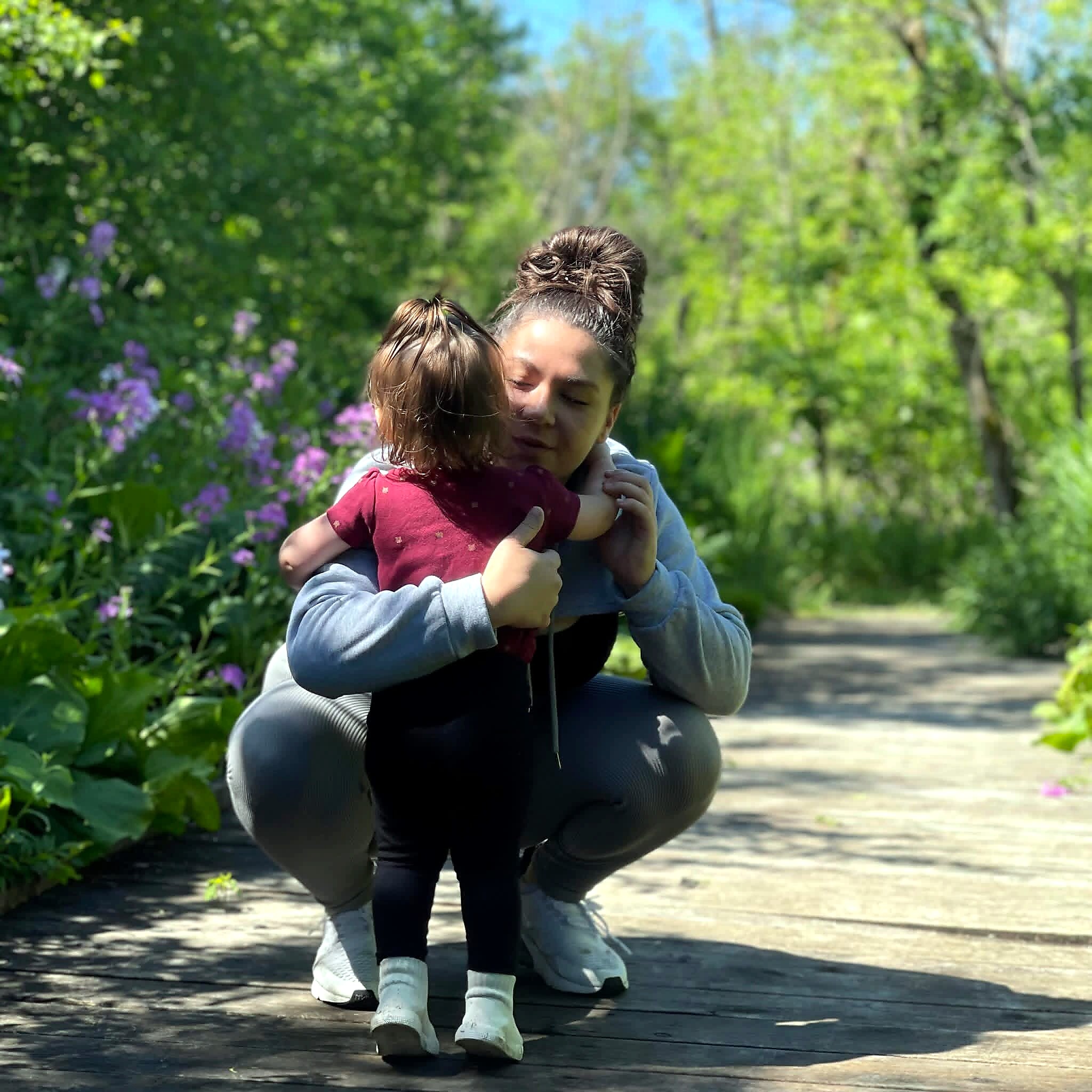Two people embrace on a sunlit wooden path surrounded by green plants and purple flowers in a garden.