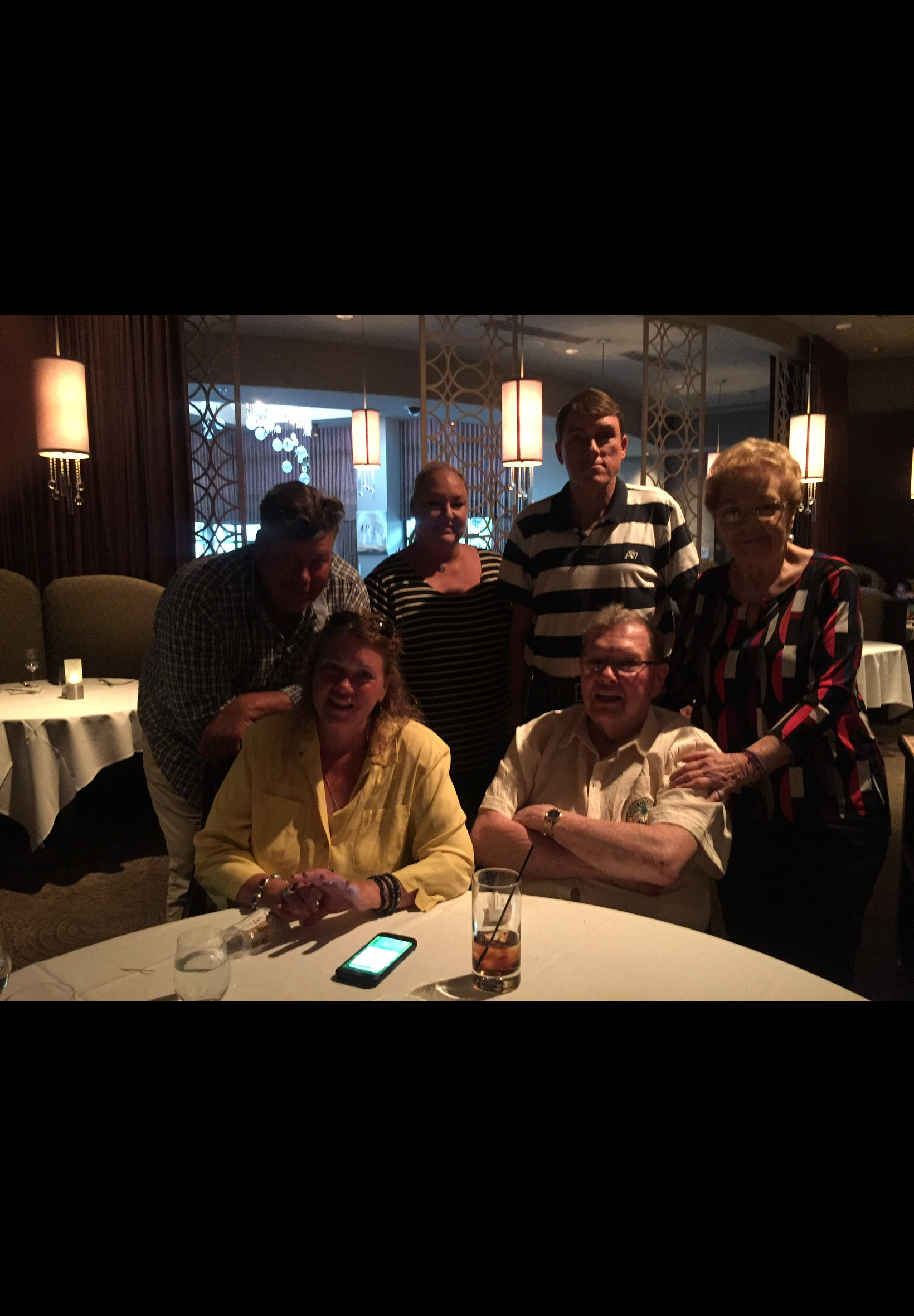 Group of adults posing for a photo around a round table in a dimly lit restaurant, with drinks and a smartphone visible on the table.