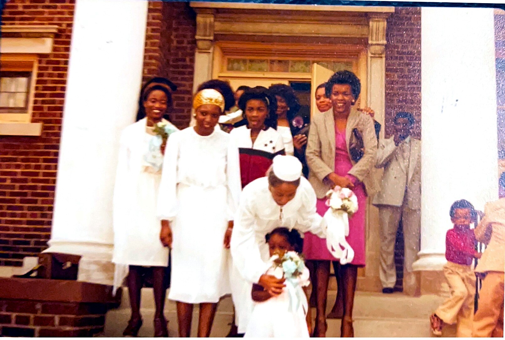 Wedding party posing on brick steps: bride in white with a gold head wrap, bridesmaids in white, and guests in formal attire; flower girl nearby.