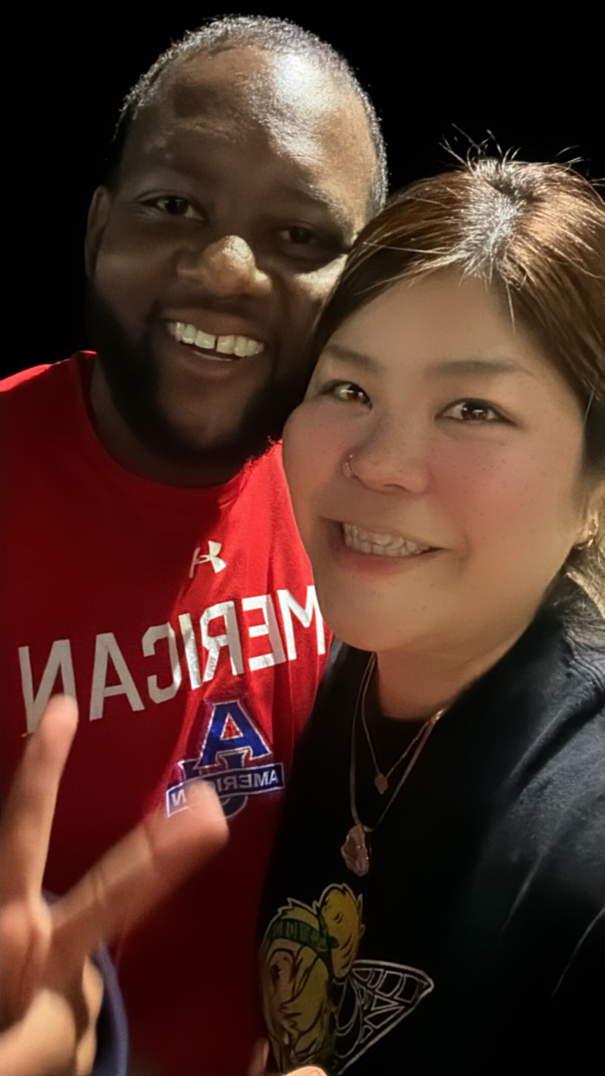 Close-up selfie of a smiling man in a red shirt and a smiling woman in a black shirt against a dark background, cropped to faces and shoulders.