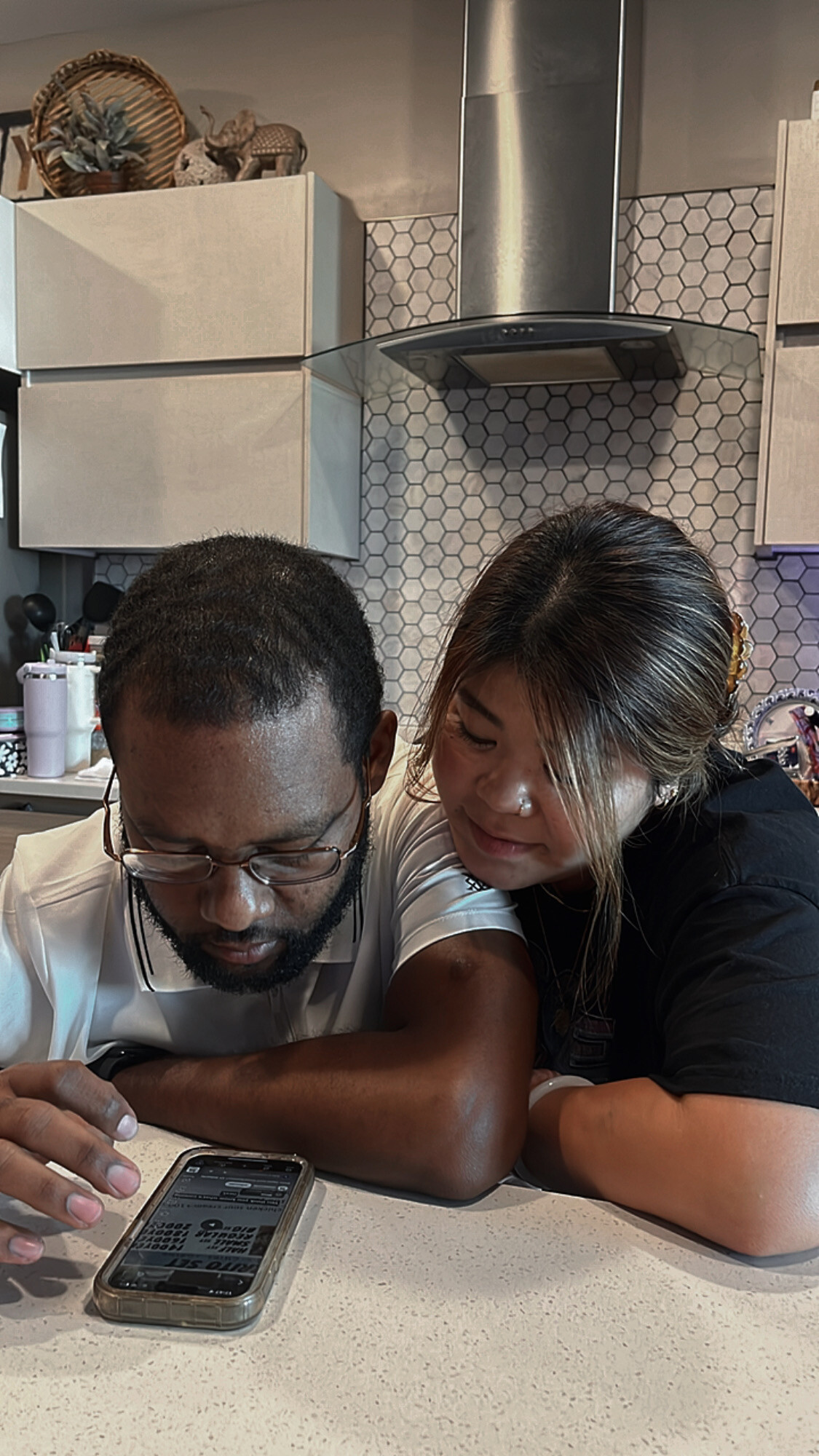 Two people leaning over a kitchen counter, looking at a smartphone together.