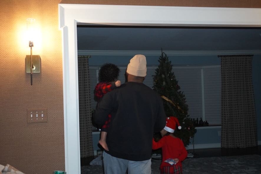 Man wearing a beanie holds a small child while a second child in a Santa hat stands by a Christmas tree in a dim living room.