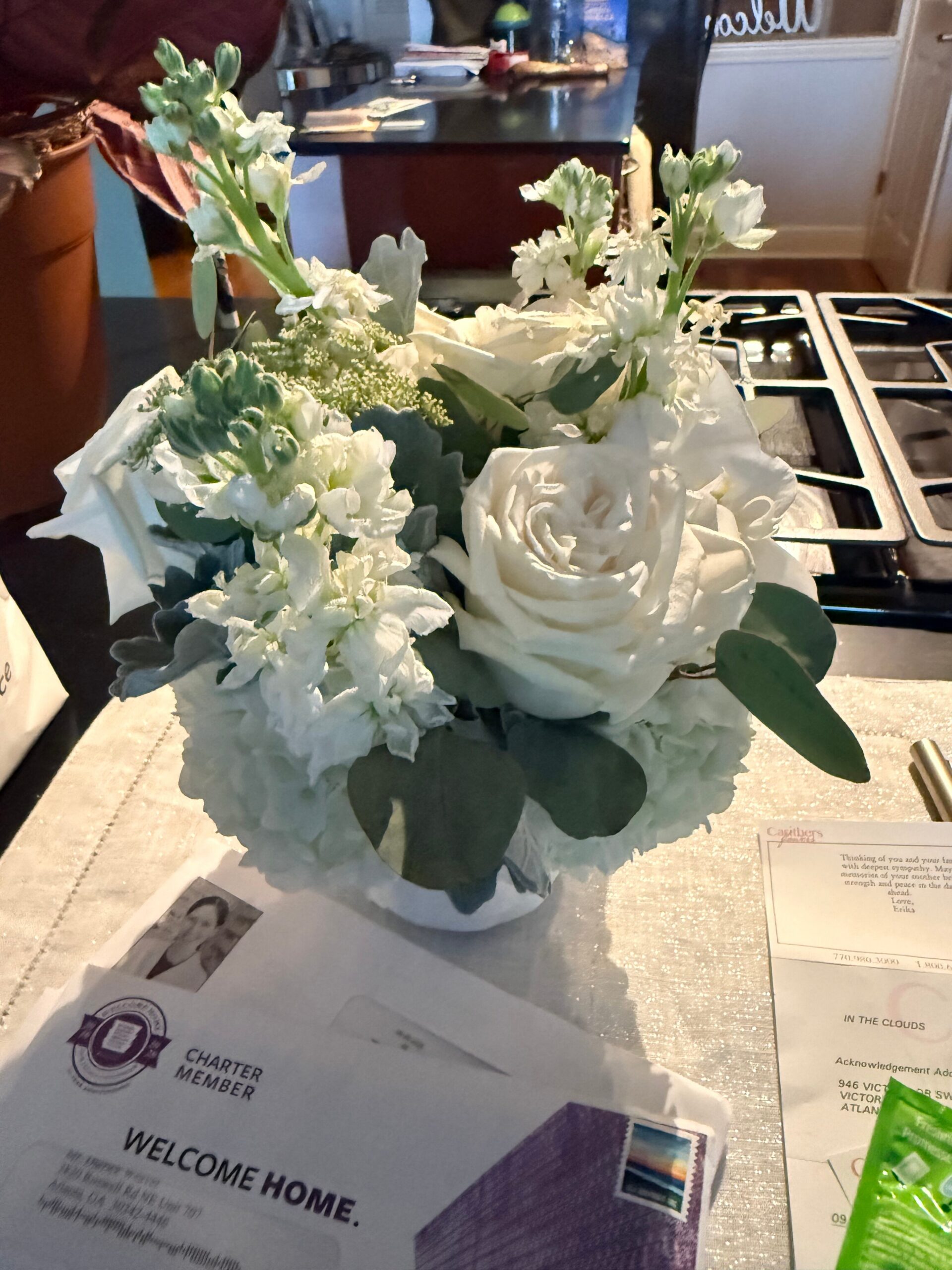 White bouquet with roses and pale greenery in vase on a kitchen or desk surface, with papers nearby and a welcome home card nearby.