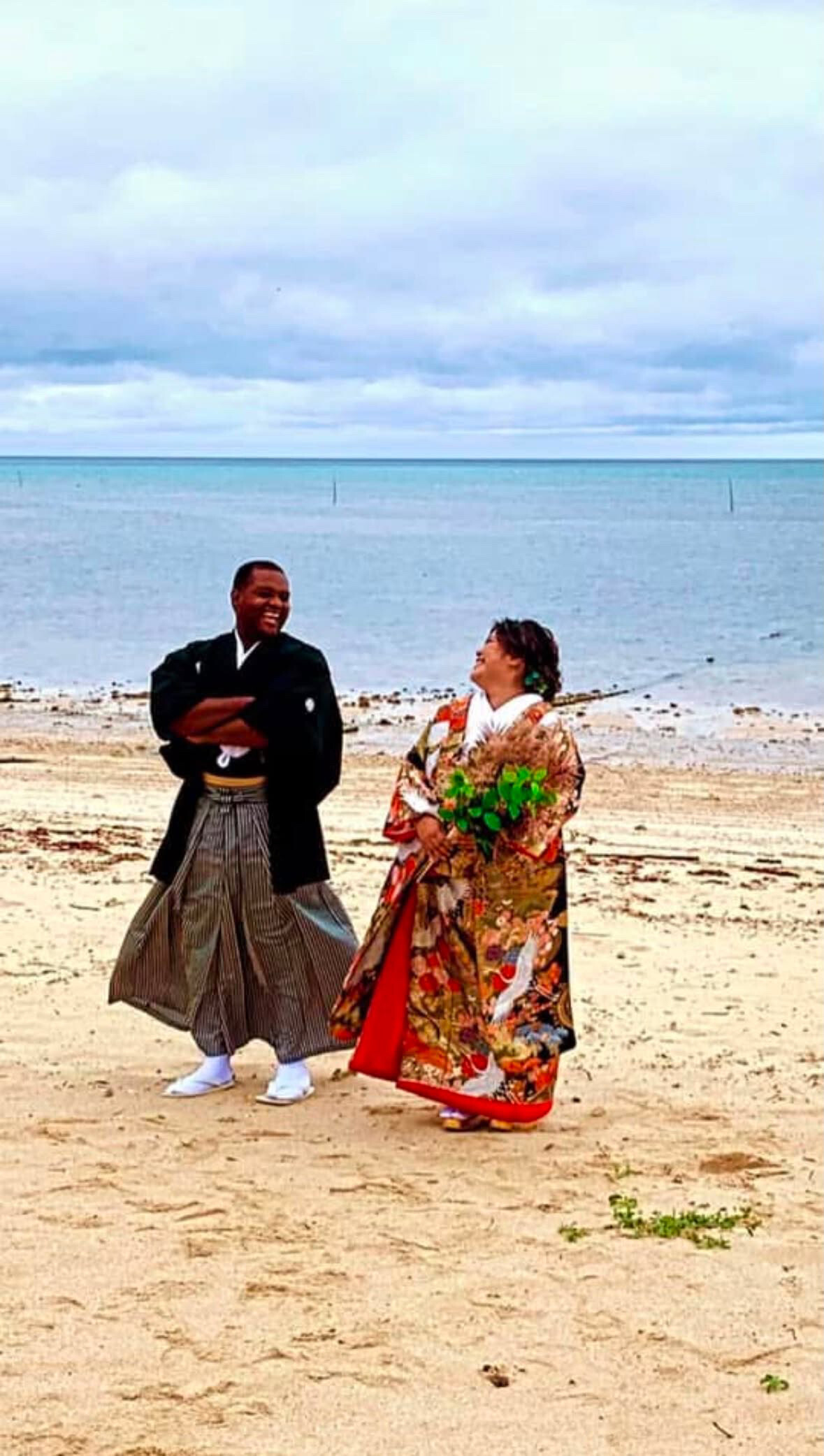A couple wearing traditional Japanese kimonos walking along a sandy beach with the sea in the background, smiling at each other.