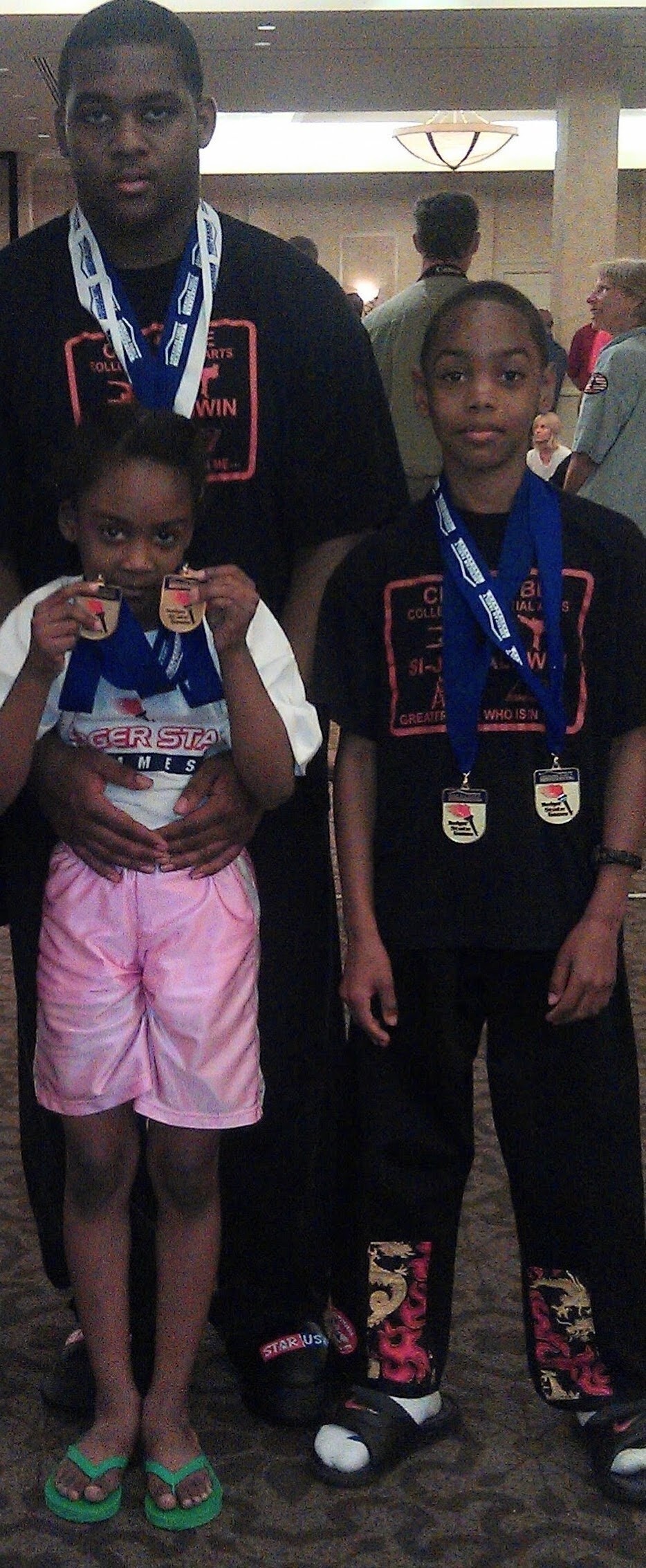 Three children and an adult posing indoors with medals around their necks; girl in white shirt and pink shorts on left, boy in black with dragon pants on right, man behind them wearing a black shirt and blue lanyard medals.