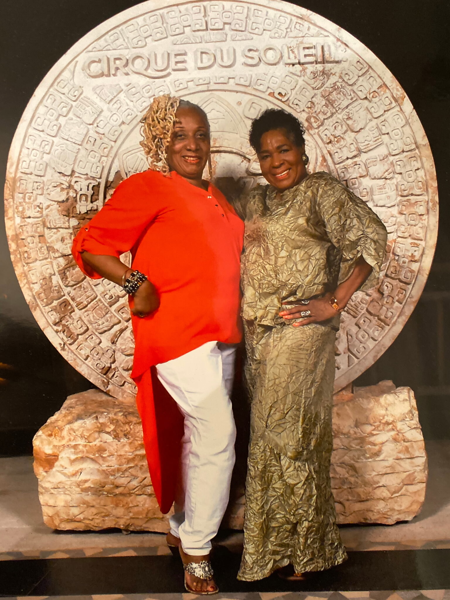 Two women smiling and posing in front of a large circular stone backdrop with Cirque du Soleil text at the top.