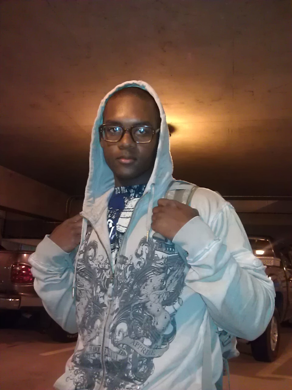 Young man wearing a light hoodie and glasses, carrying a backpack in a dim parking garage at night.