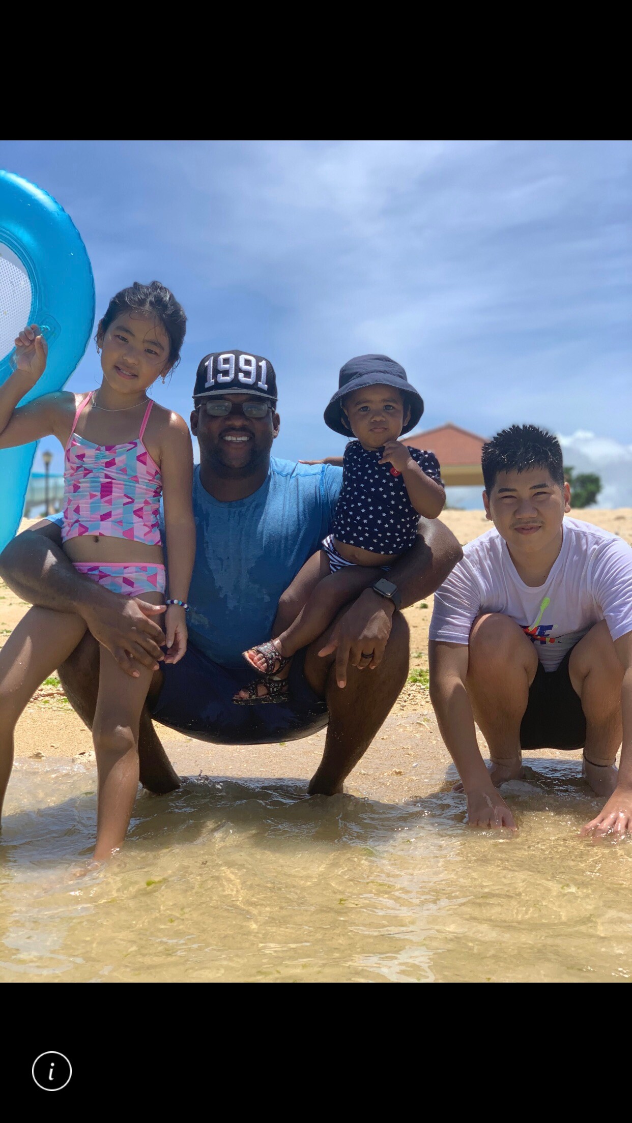Family at the beach: man kneels in shallow water with his daughter on one side and a toddler on his other knee, all smiling as waves wash in.