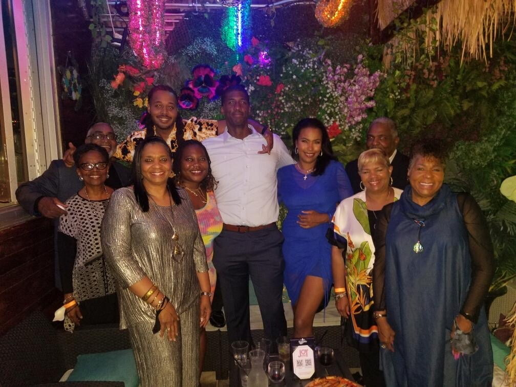 Group of ten adults posing together at a festive indoor party with colorful lights and a lush floral backdrop, smiling for the photo.