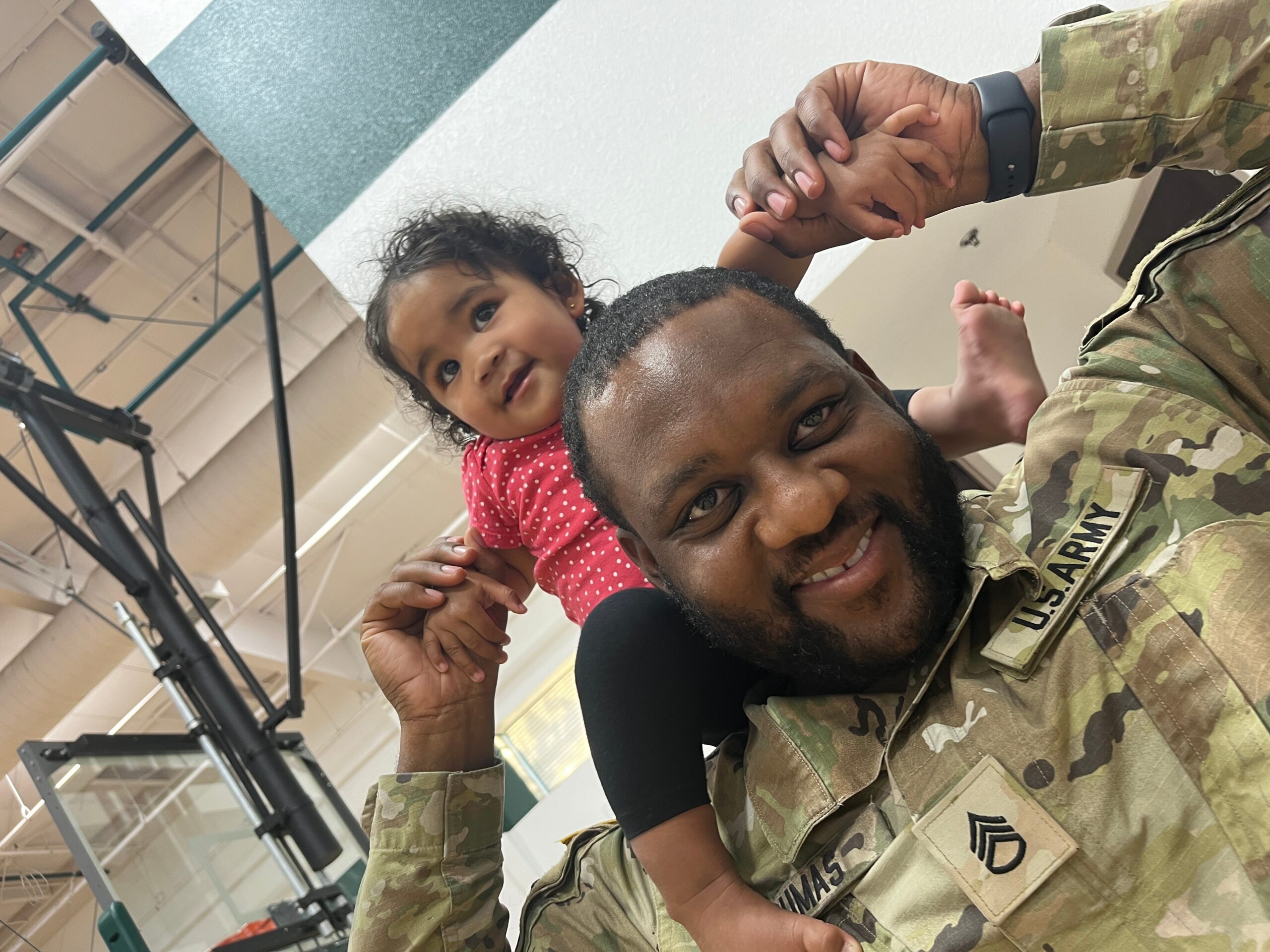 A person in camouflage military uniform holding hands with a smiling toddler perched on their shoulders indoors.