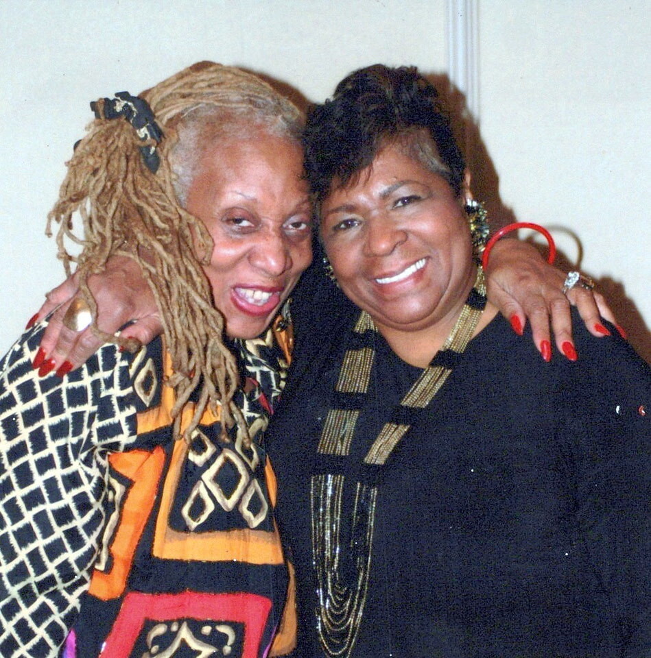 Two women smiling with arms around each other, one in a bold geometric dress with dreadlocks and red nails, the other in black with gold jewelry and short hair.