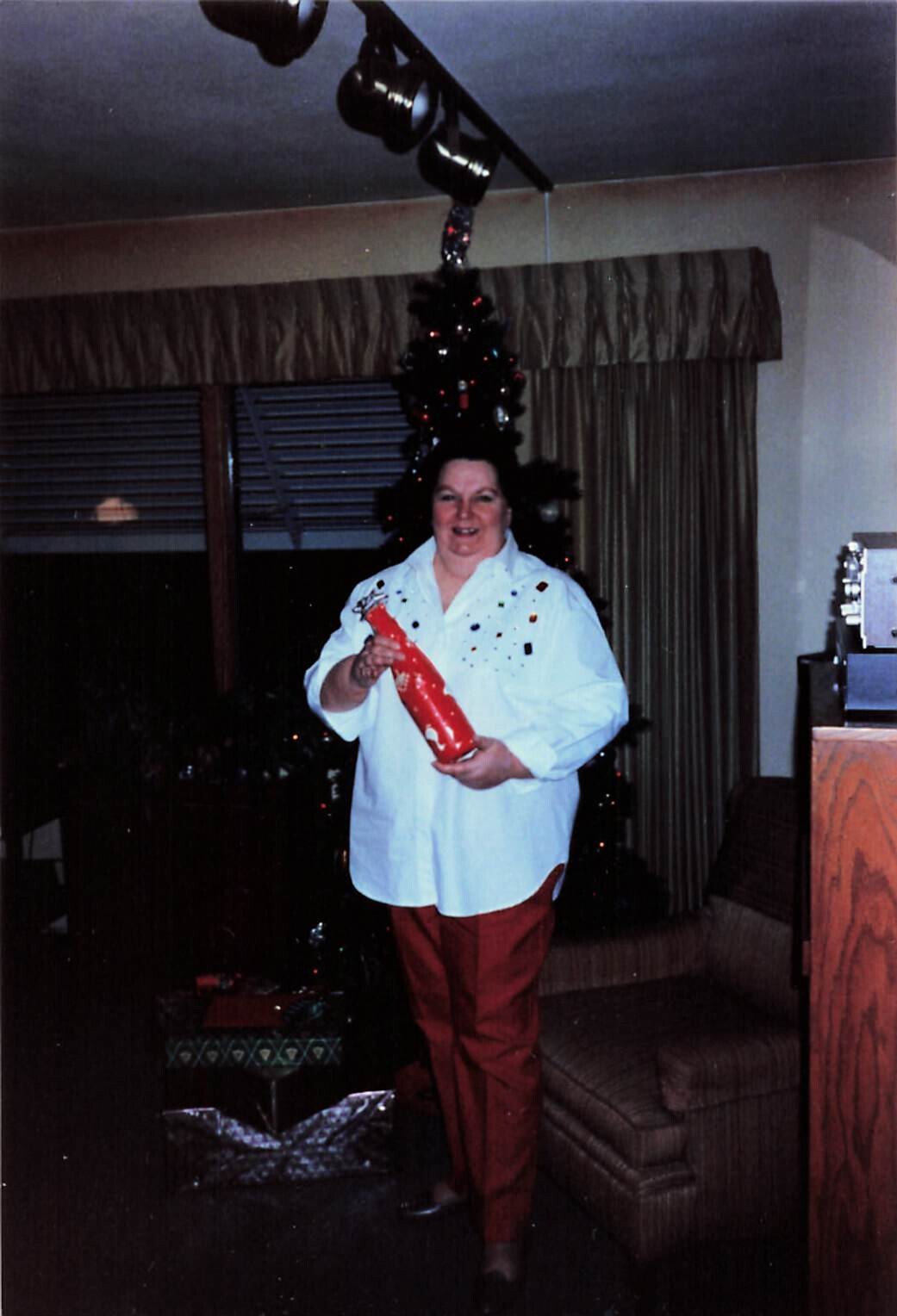 Woman standing in a living room, holding a bright red gift wrap or tube, with a Christmas tree behind her and festive decorations in view.