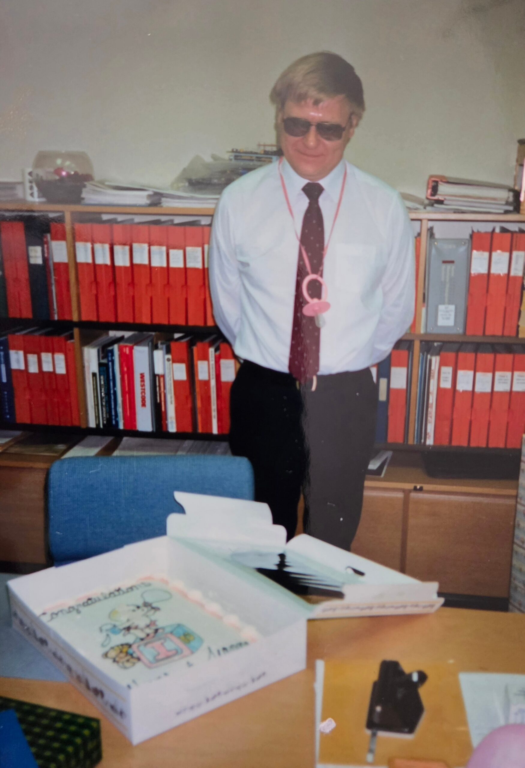 Man in a white shirt and tie with sunglasses, standing in front of a shelf filled with red binders in an office.